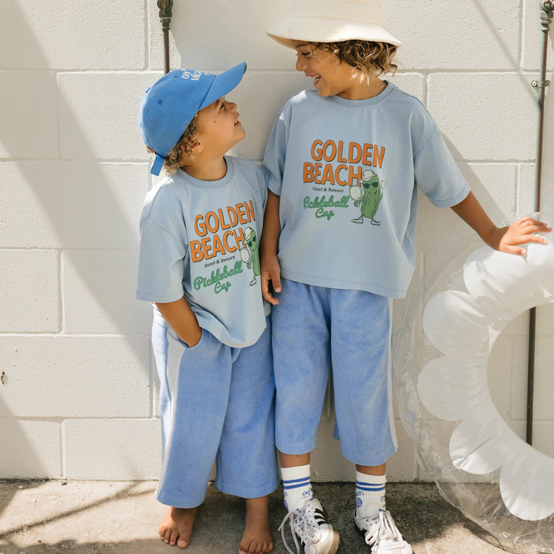 Two children stand against a white wall, both dressed in relaxed fit light blue outfits and caps from GOLDEN CHILDREN. Their Golden Beach Mid Sleeve Tees in Palm Blue read "Golden Beach." They smile at each other, with one holding a white inflatable ring as sunlight casts gentle shadows on the ground.