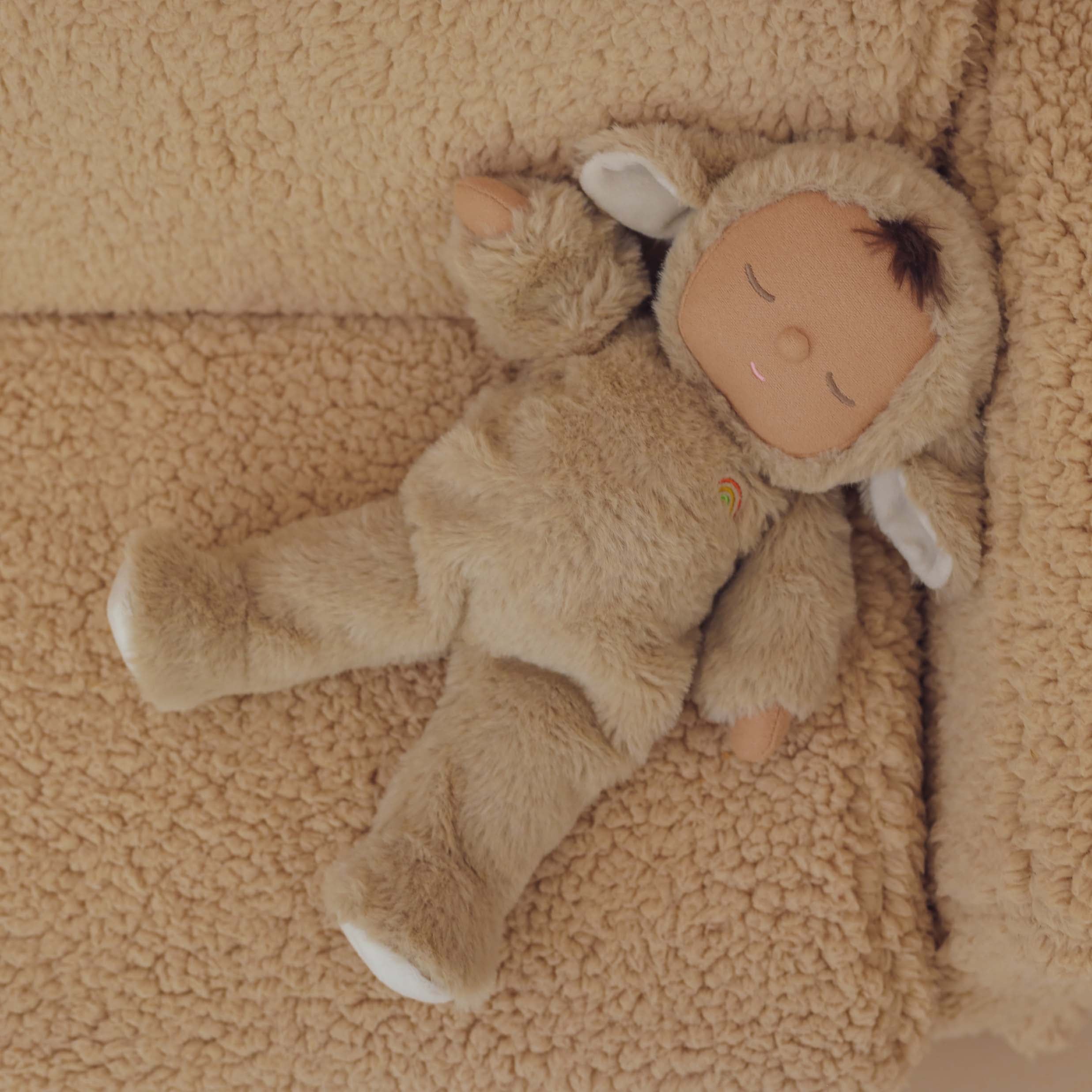 A Cozy Dinkum Lamby Pip from OLLI ELLA, dressed in a fuzzy beige lamb costume with closed eyes, is lying on a textured tan blanket. This snuggly collectible features a small rainbow embroidered on its chest and has white ears peeking out from the hood.