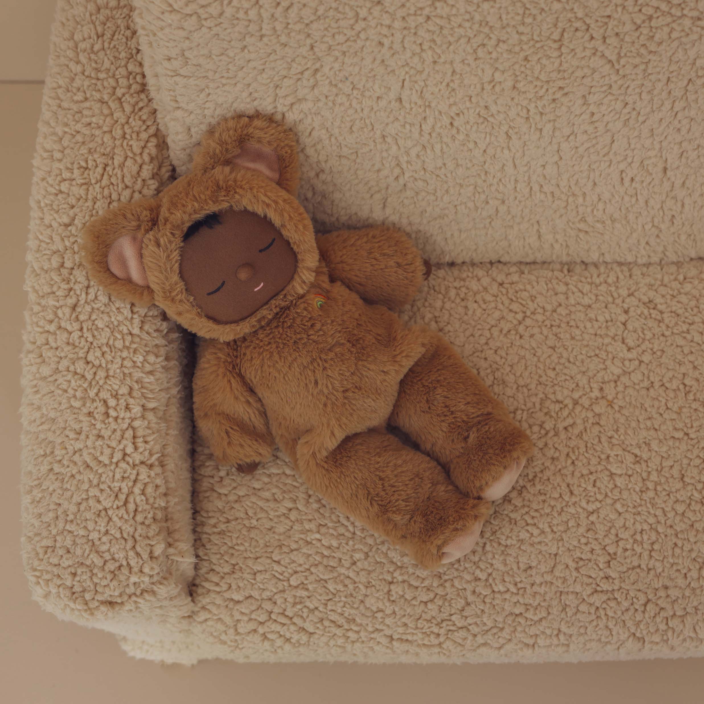 A Cozy Dinkum Teddy Mini from OLLI ELLA, resembling a baby in a brown bear costume with a hood that sports adorable bear ears, lies on its back on a textured beige cushion, appearing peacefully asleep with closed eyes. This snuggly collectible is perfect for adding to your collection.