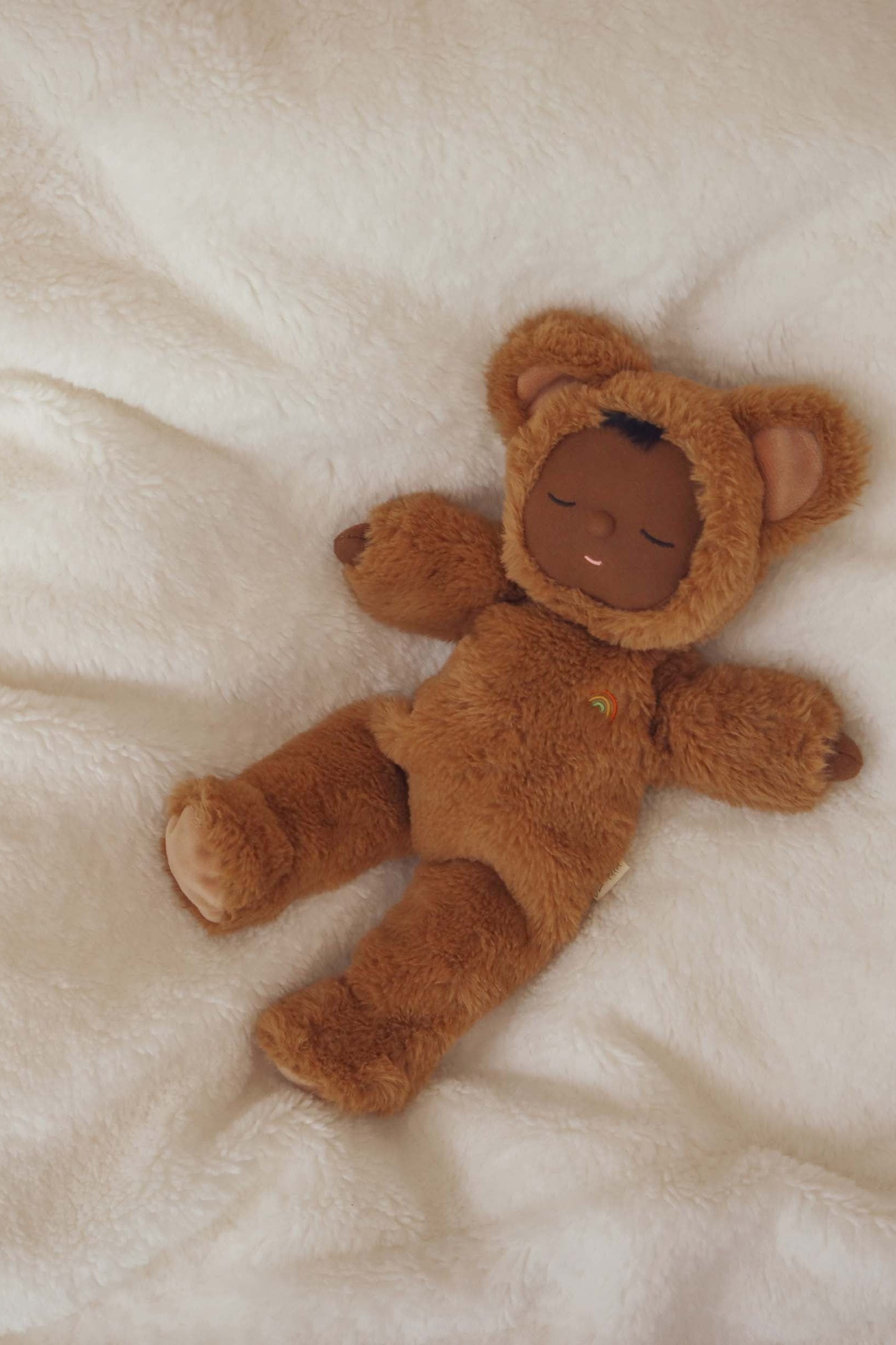 A Cozy Dinkum Teddy Mini from OLLI ELLA, with brown skin and dressed in a fuzzy bear costume featuring bear ears, lies on a soft, cream-colored blanket. The doll's eyes are closed, giving the appearance of sleeping, and there is a small rainbow emblem on its snuggly collectible costume.