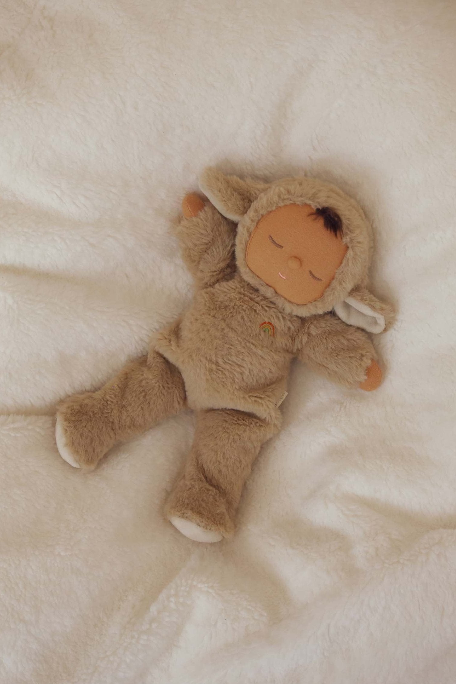 A charming collectible, the OLLI ELLA Cozy Dinkum Lamby Pip, is attired in a fuzzy beige lamb costume complete with a hood featuring small ears and lies on a soft, white blanket. With its eyes gently closed, this plush baby doll evokes a serene sleeping appearance.