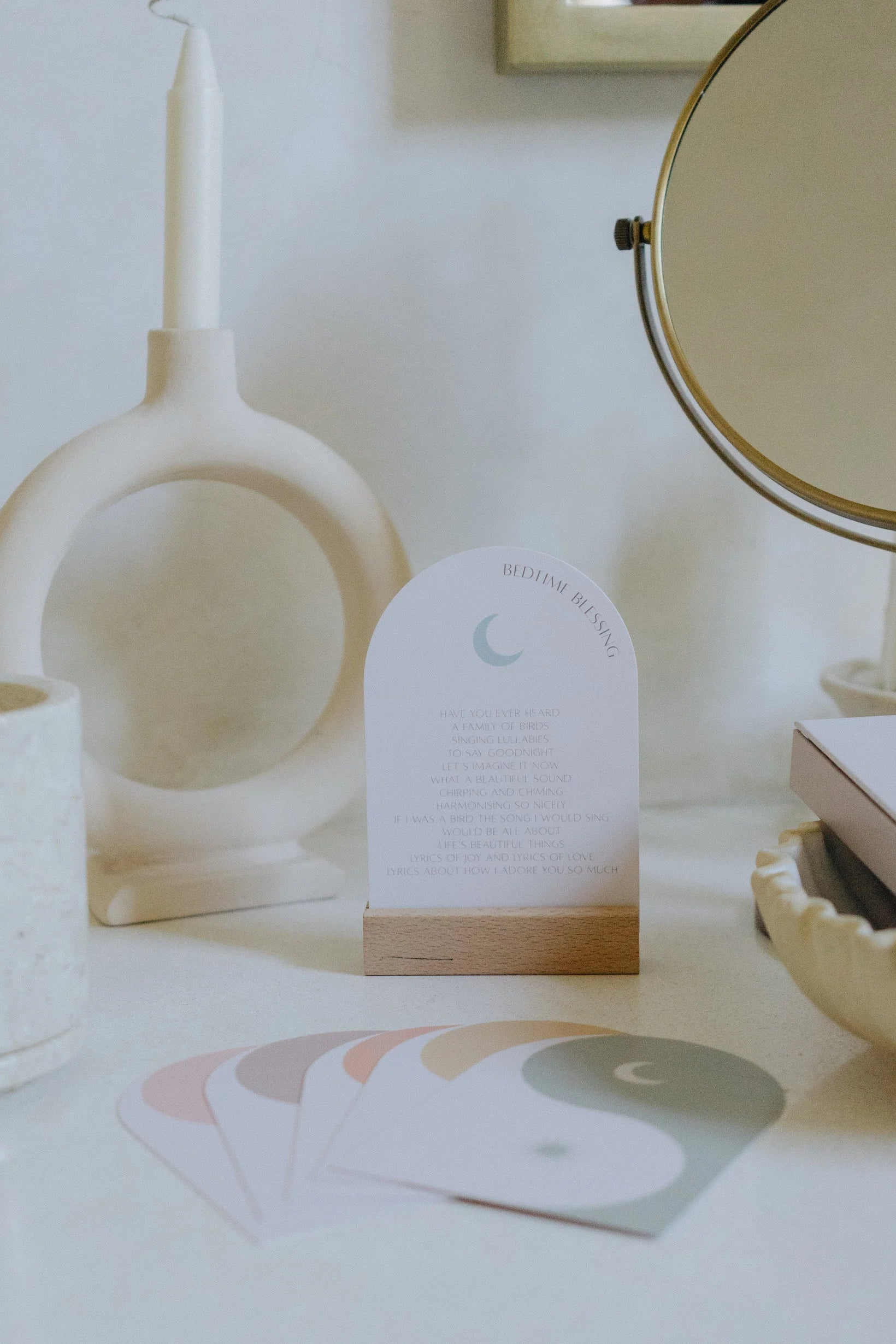 A small card from the *Bedtime Blessings* by *HENY AND HAZE* is displayed on a wooden stand. The setup includes a round mirror, a modern candle holder with a lit white candle, and a concrete pot. Additionally, five pastel-colored affirmation cards from the *Bedtime Blessings Deck* are laid out in front.