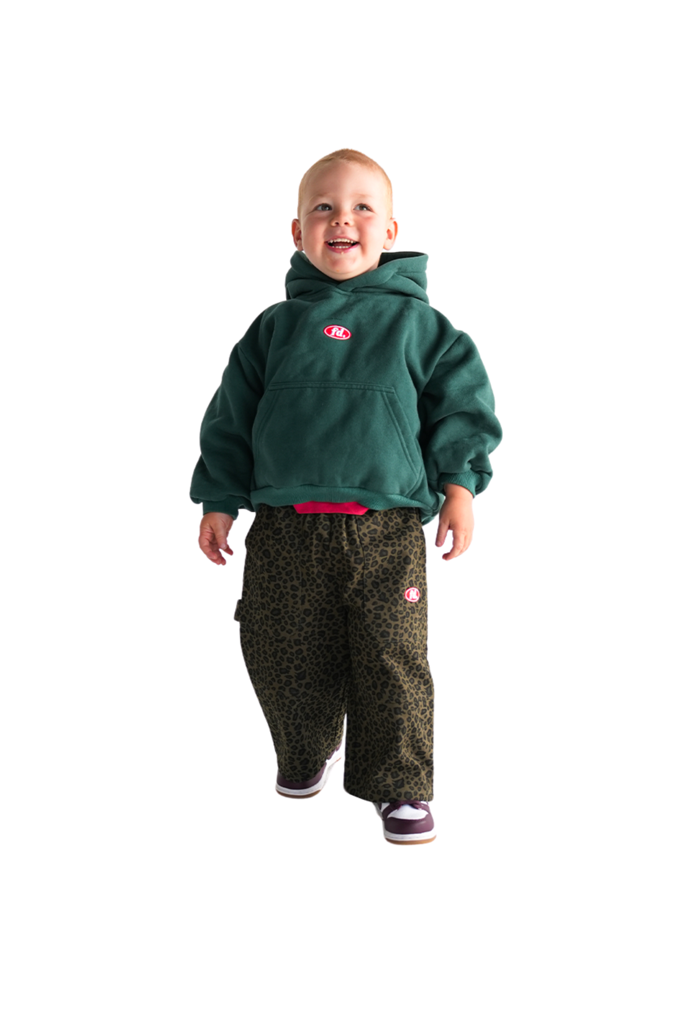 A cheerful toddler sports a green hoodie with a red logo and confidently models FRANCO'S DAD ~ PRE-ORDER Leopard Cargos, featuring an oversized fit and stretch waistband. The child pairs them with dark sneakers that have purple accents, all on a plain white backdrop.