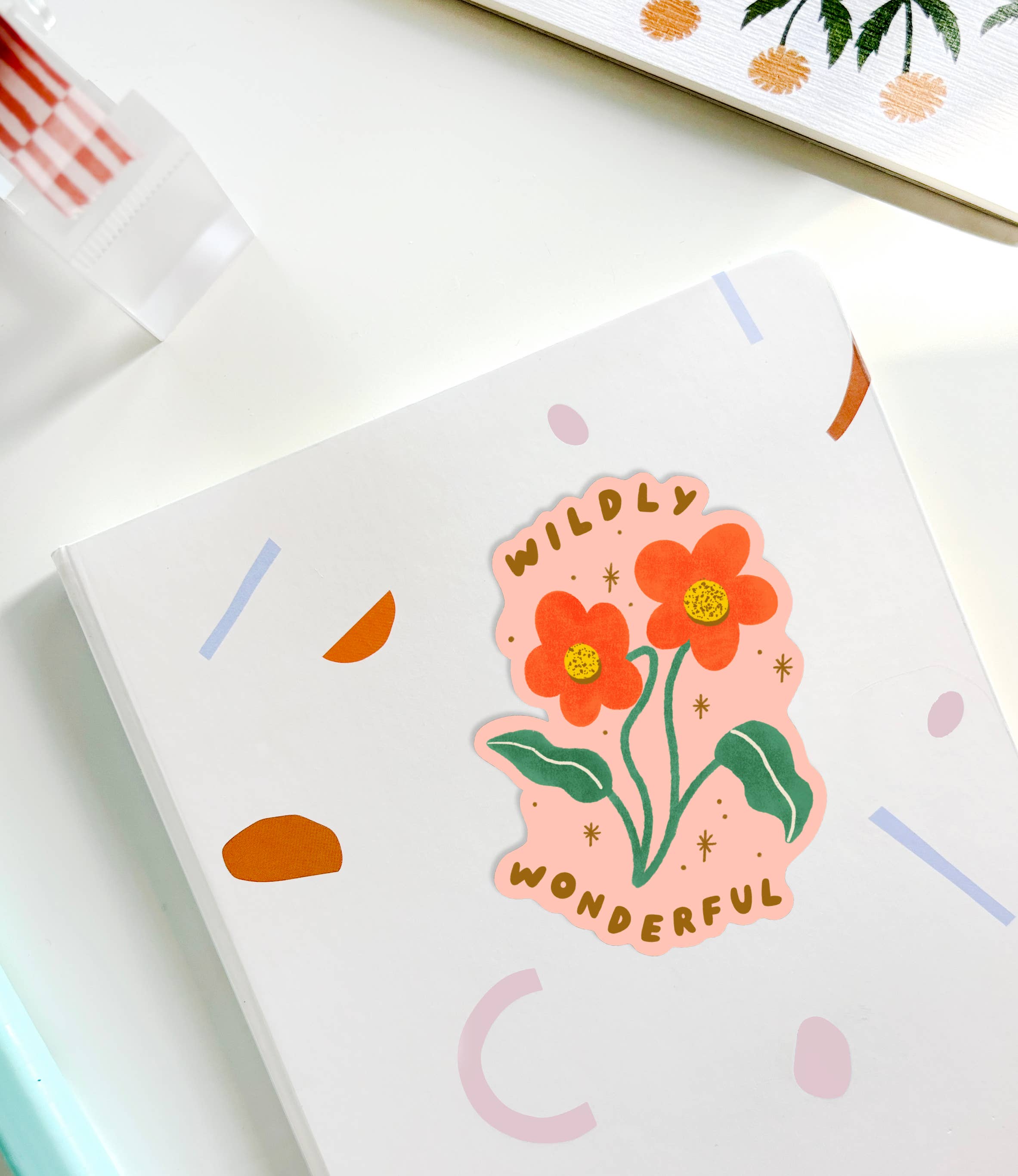 A white notebook adorned with LAUREN SISSONS STUDIO's Wildly Wonderful Sticker, showcasing an orange and red flower with the inspiring words "Wildly Wonderful." This charming sticker enhances the scene, which also includes glimpses of a plastic fork, a plant illustration, and a mint green object nearby.