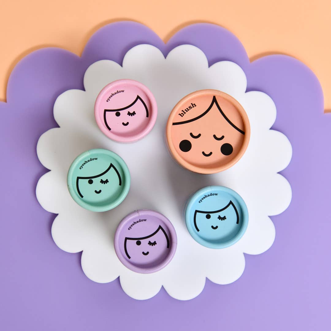 Skincare containers with cartoon faces set against pastel backgrounds include two in pink, two in turquoise, and one in lavender. The largest container at the center is labeled "Kids Eco Natural Pressed Eyeshadow & Blush" by no nasties kids, featuring cruelty-free and natural ingredients. These containers are displayed on a scalloped white paper resting on purple and peach surfaces.