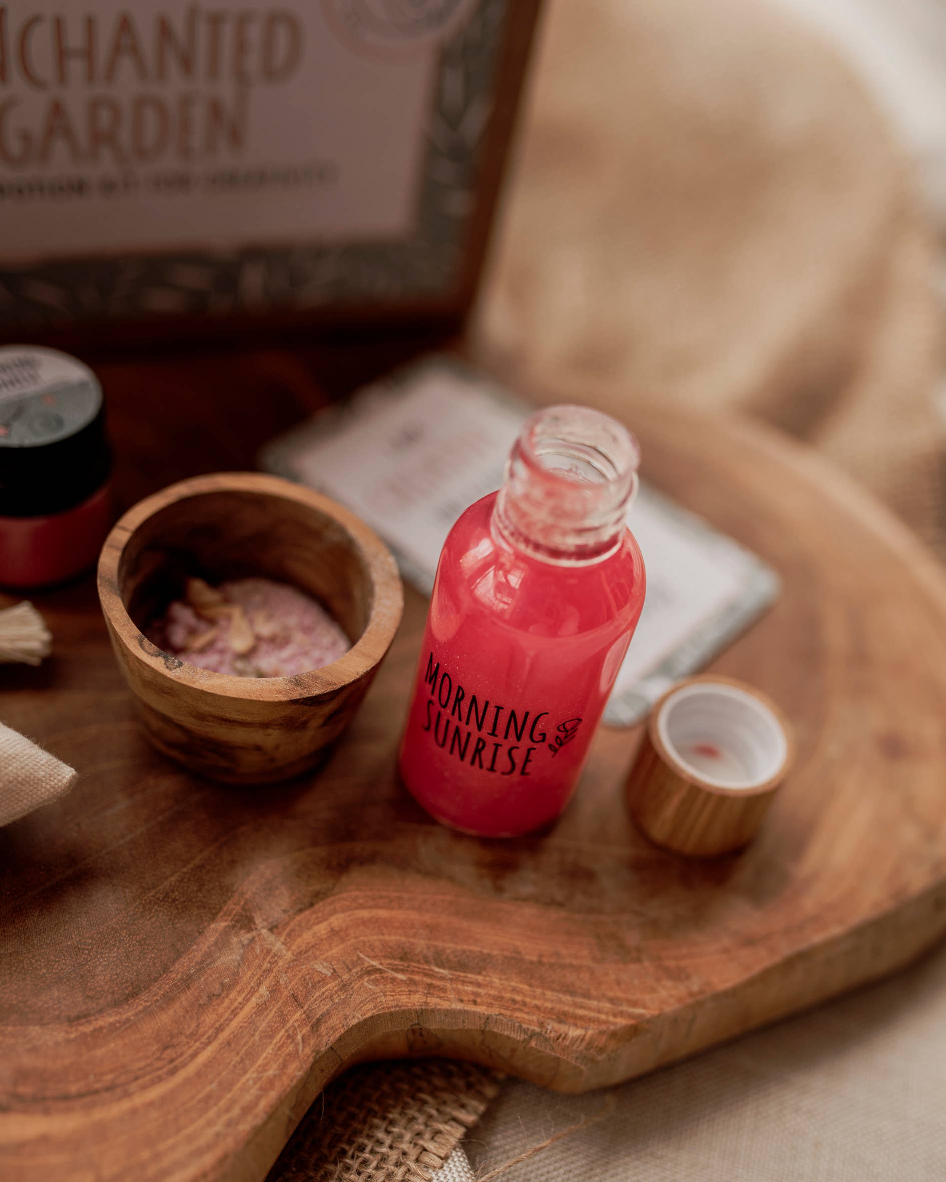 A petite glass bottle labeled "Morning Sunrise" containing a pink liquid rests on a wooden surface as part of THE LITTLE POTION CO's enchanting MINI Enchanted Garden Potion Kit. The bottle's cap is off and sits beside it. Nearby, there is a wooden bowl filled with pink bath salts. In the background, a box with partial text can be seen.