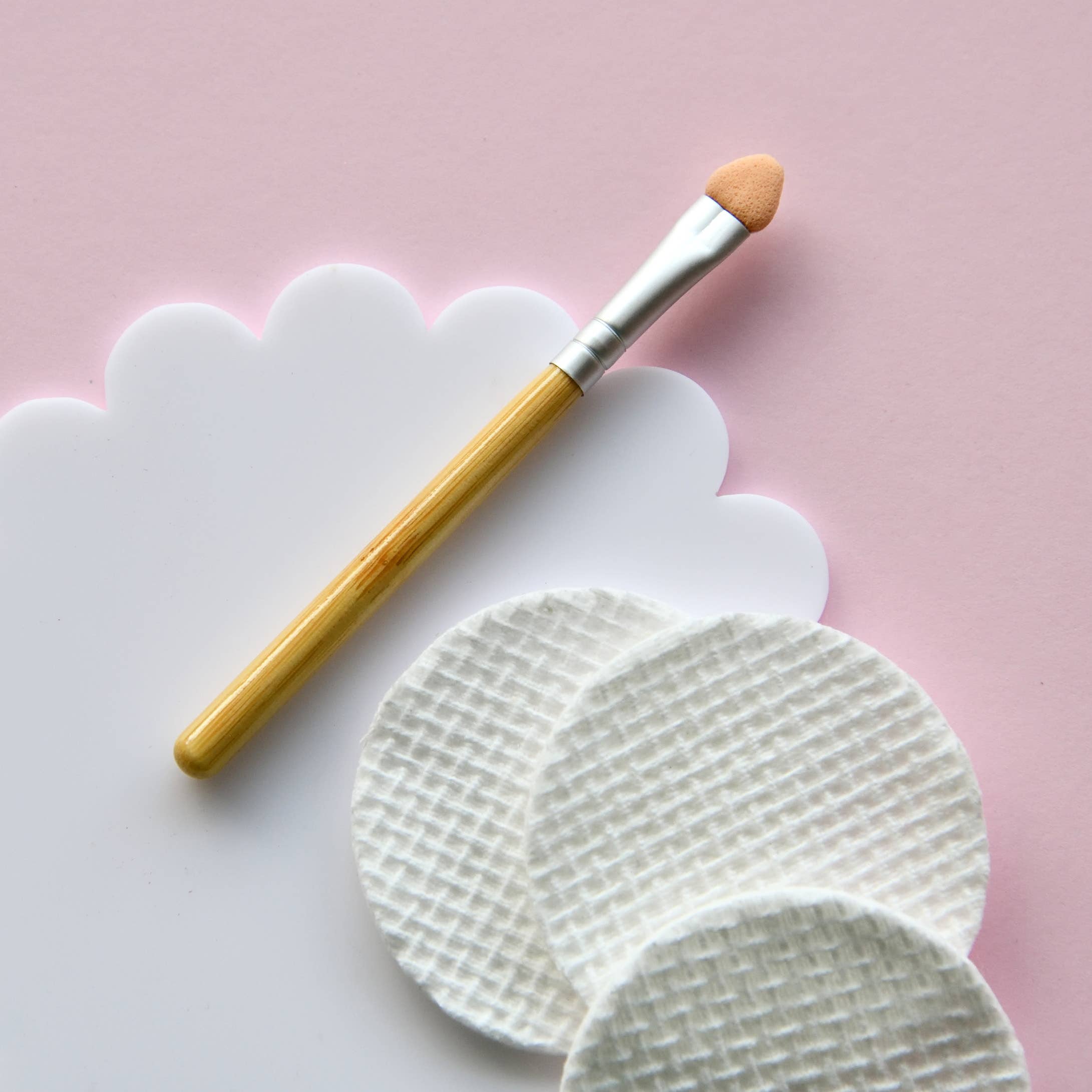 A makeup applicator from the Kids Eco Natural Pressed Eyeshadow & Blush by no nasties kids, featuring a wooden handle, is placed next to three textured cotton pads on a pink and white scalloped background. This cruelty-free tool, made with natural ingredients, ensures your beauty routine is gentle and mindful.