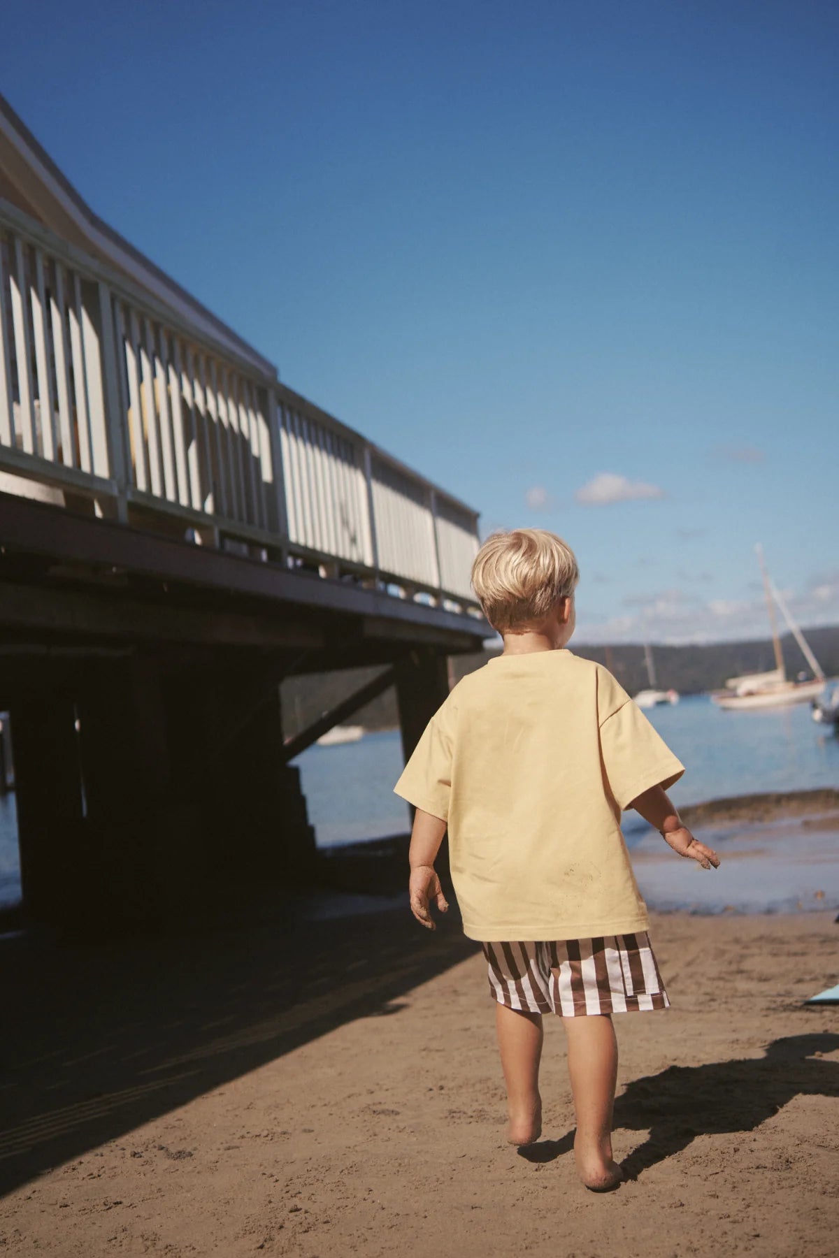 A young child with blonde hair, in an oversized Little Lou Tee Pomello from LITTLE THE LABEL and checkered shorts, walks barefoot on a sandy beach toward a wooden boardwalk. Boats float on a calm blue sea under a clear sky in the background.