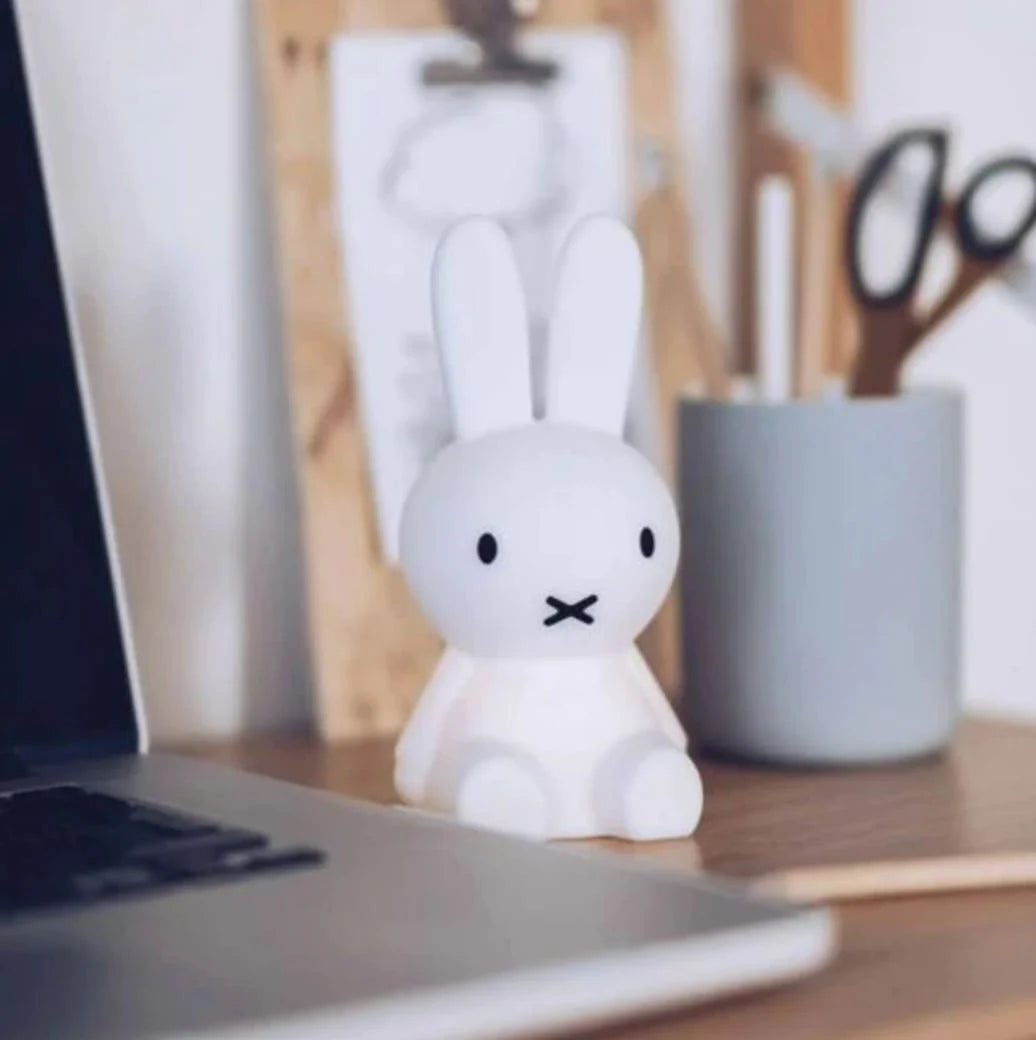 A small, white "Bundle of Light - Miffy" figurine with black eyes and a cross for a mouth from MR MARIA sits on a wooden desk near an open laptop, a pot of scissors, and a clipboard. The background is slightly blurred, highlighting the Miffy as the focal point—a charming addition to any gift display items.