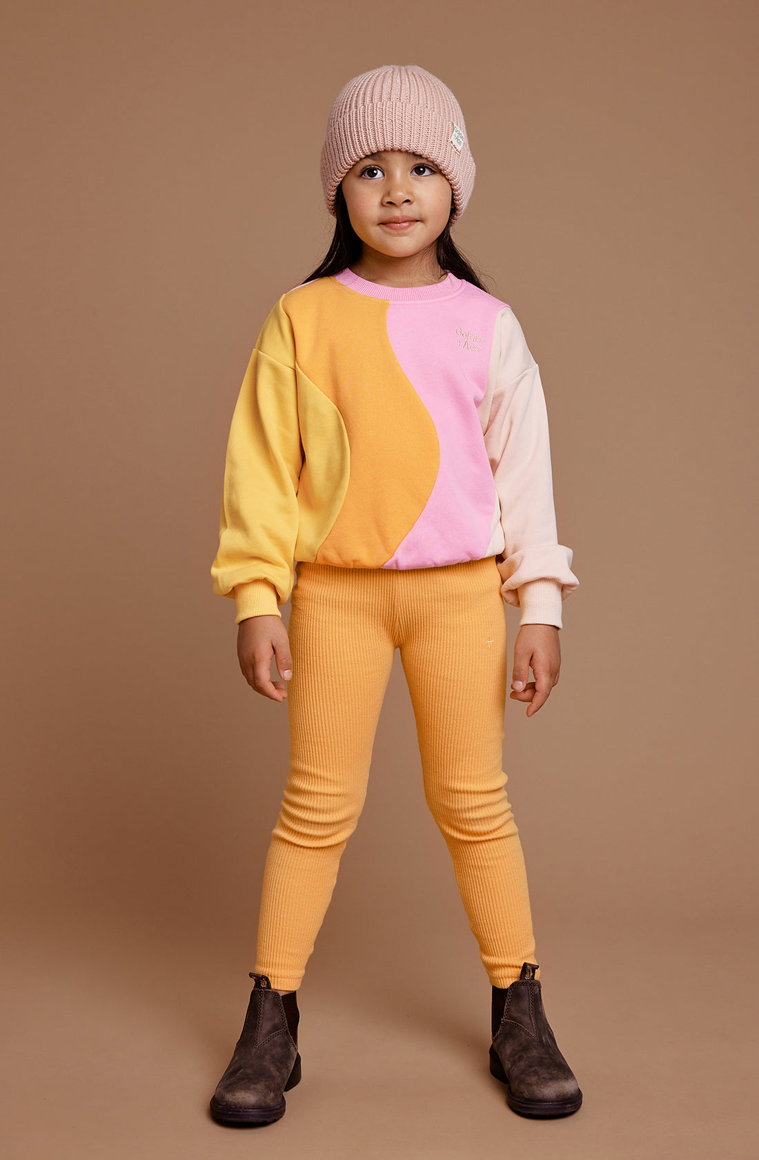 A young girl wearing the Rio Wave sweater in pink and gold multi design by Goldie + Ace. She also wearing a pastel pink beanie and orange tights with brown boots.