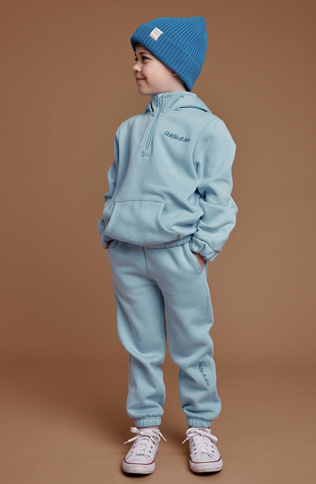 A young child stands against a plain brown background, wearing a light blue hooded sweatshirt with a generous fit, matching jogging pants, white sneakers, and the "Wool Beanie Lake" by GOLDIE + ACE. The child has their hands in their pockets and is looking to the side with a slight smile.