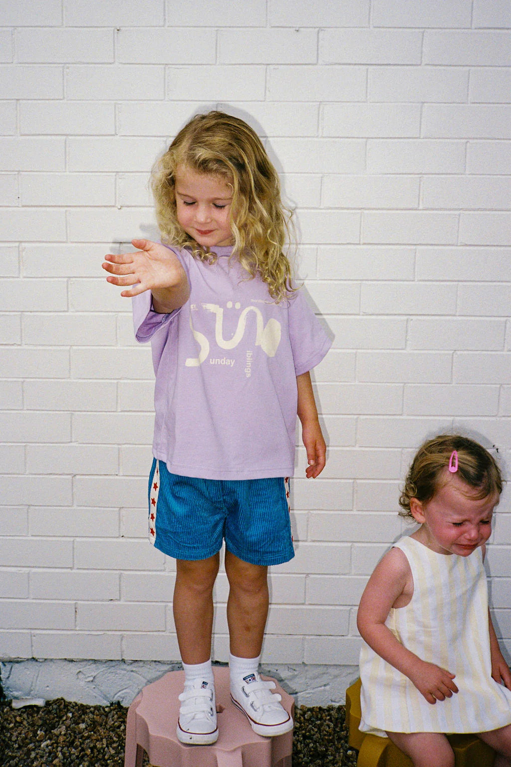 A young child with curly hair stands on a stool, wearing a SUNDAY SIBLINGS Il Sole Tee in Purple/Cream and blue shorts. Beside them, another child in a striped dress sits against a white brick wall, looking upset.