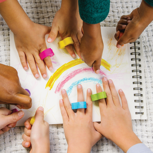 A bunch of kids hands with OOLY heart ring crayons on their fingers drawing on a piece of paper.