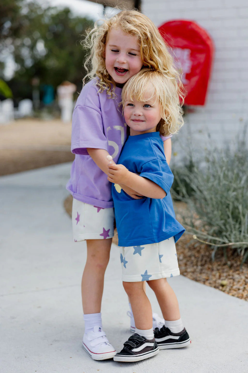 Two curly-haired kids embrace outdoors, one in purple and the other in blue, wearing matching Sunday Siblings Kiddo Shorts Purple with star prints. A red object and greenery can be seen in the sunny background.