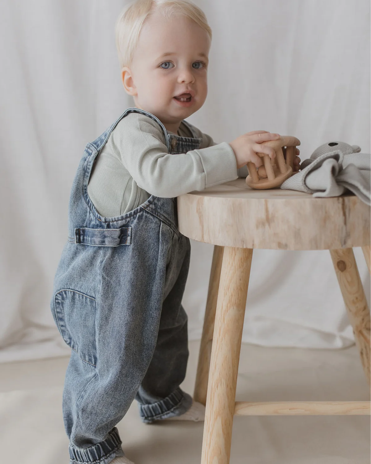 A toddler with blonde hair stands next to a wooden table, wearing a light gray long-sleeved shirt and SUSUKOSHI's Denim Overalls Sky Blue, featuring adjustable shoulder straps. They hold a wooden toy while a gray cloth is draped on the table. The overalls are crafted from ethically made organic cotton, complementing the soft background of light-colored curtains.