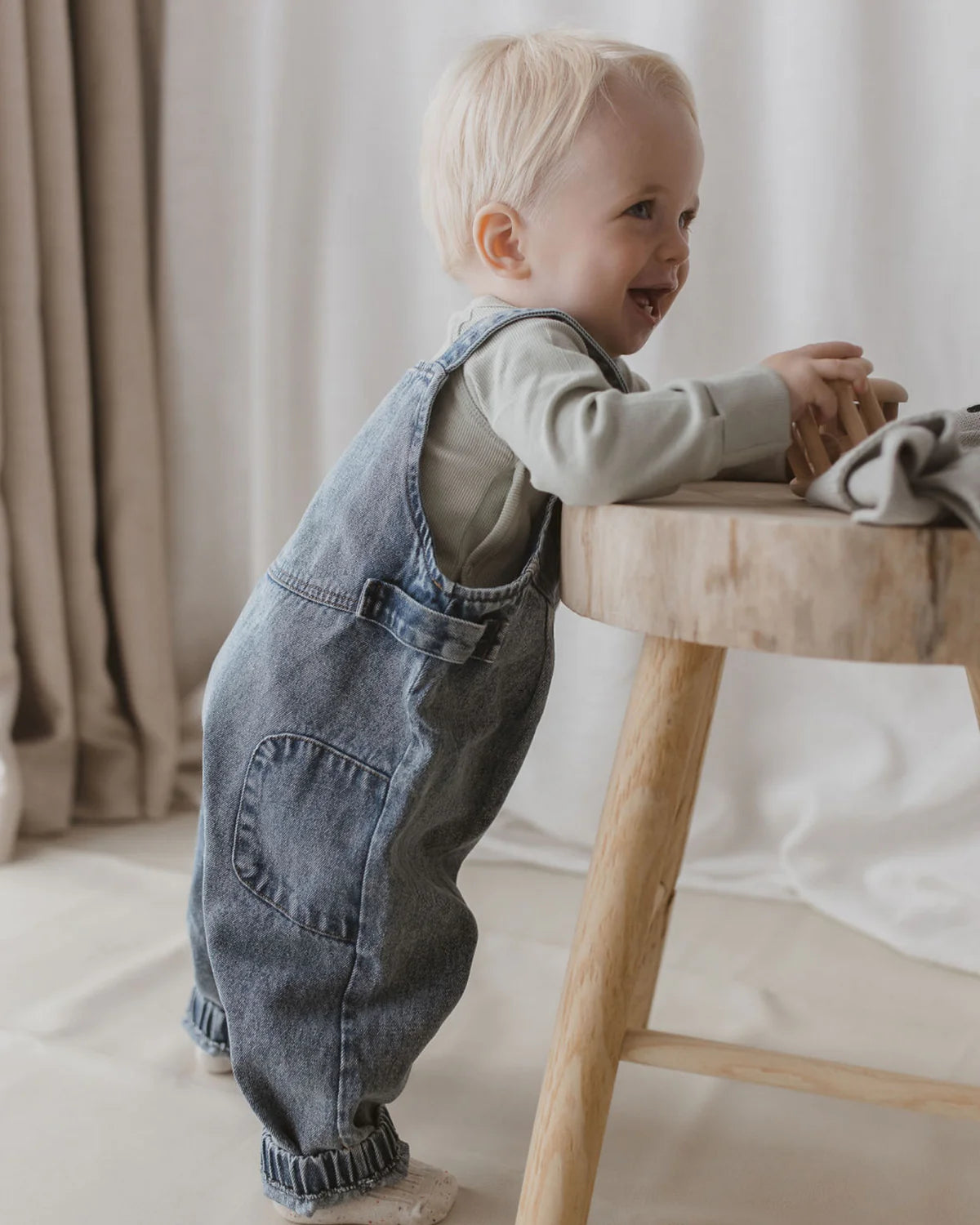 A smiling toddler with blonde hair stands while leaning against a wooden table, wearing Denim Overalls Sky Blue by SUSUKOSHI, featuring adjustable shoulder straps and paired with a long-sleeved gray shirt. The child’s outfit is made from ethically-produced organic cotton. In the background, light-colored and brown curtains softly frame the scene.