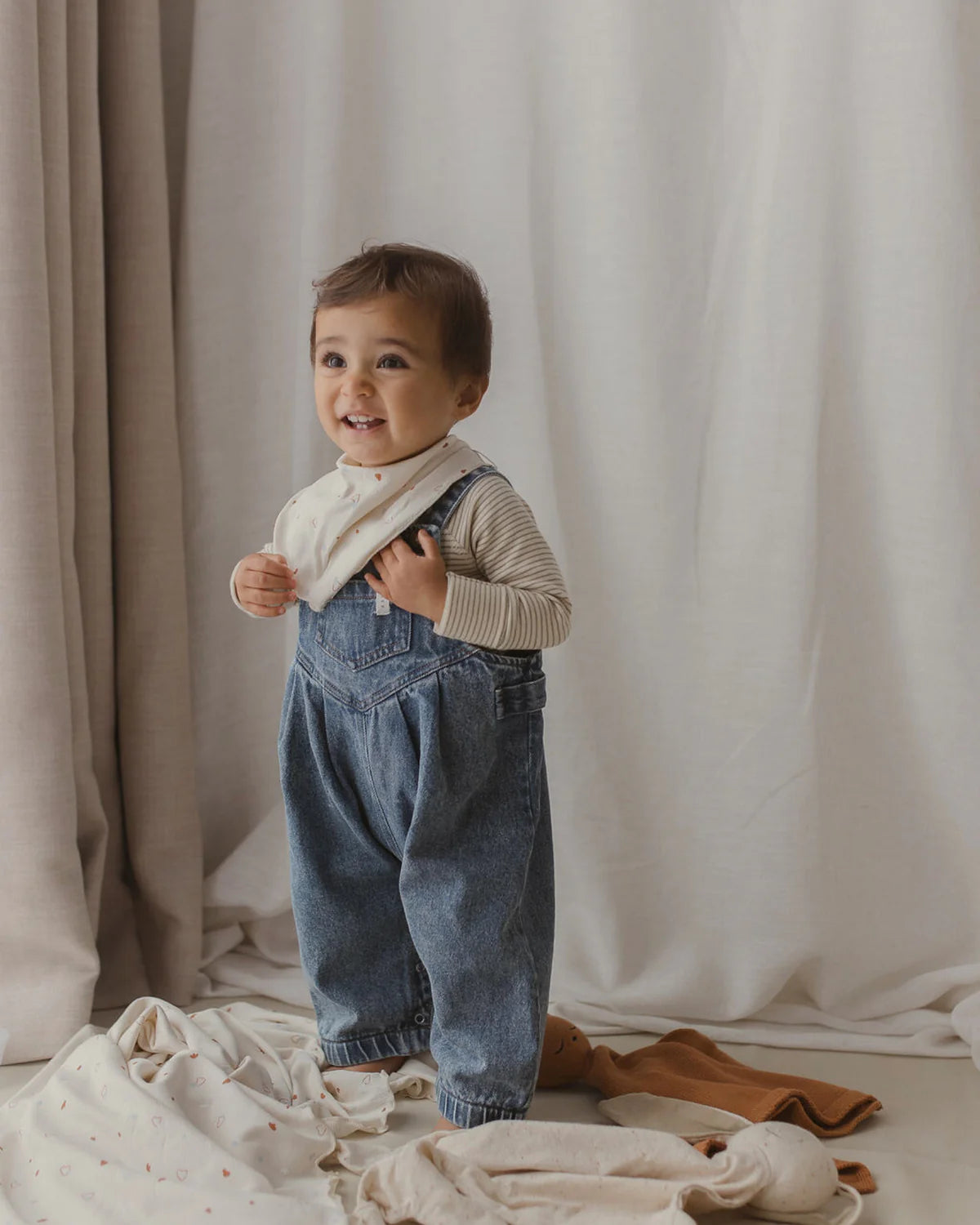 A young child stands smiling, wearing a light striped shirt and a pair of Denim Overalls Sky Blue by SUSUKOSHI, featuring adjustable shoulder straps and a white bib. These ethically made overalls perfectly match the joyful scene. The child holds the straps, standing in front of a light-colored curtain, surrounded by crumpled clothing items on the floor.