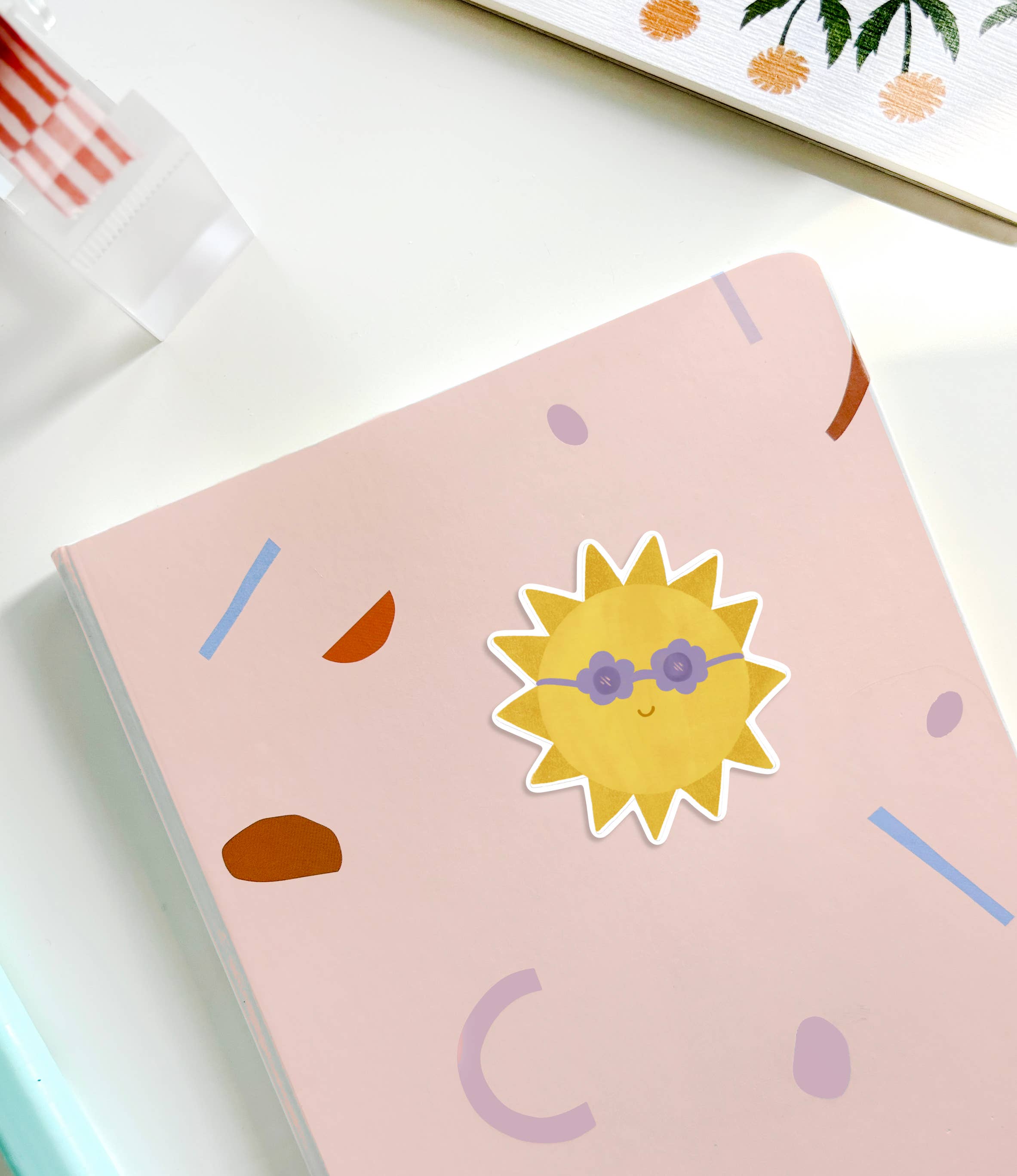 A pink notebook from LAUREN SISSONS STUDIO radiates positive vibes with a cheerful Sunshine Sticker featuring a sun wearing blue sunglasses. Abstract shapes in various colors adorn the cover, while the edge of a floral-patterned item and a red and white striped object peek out in the background.