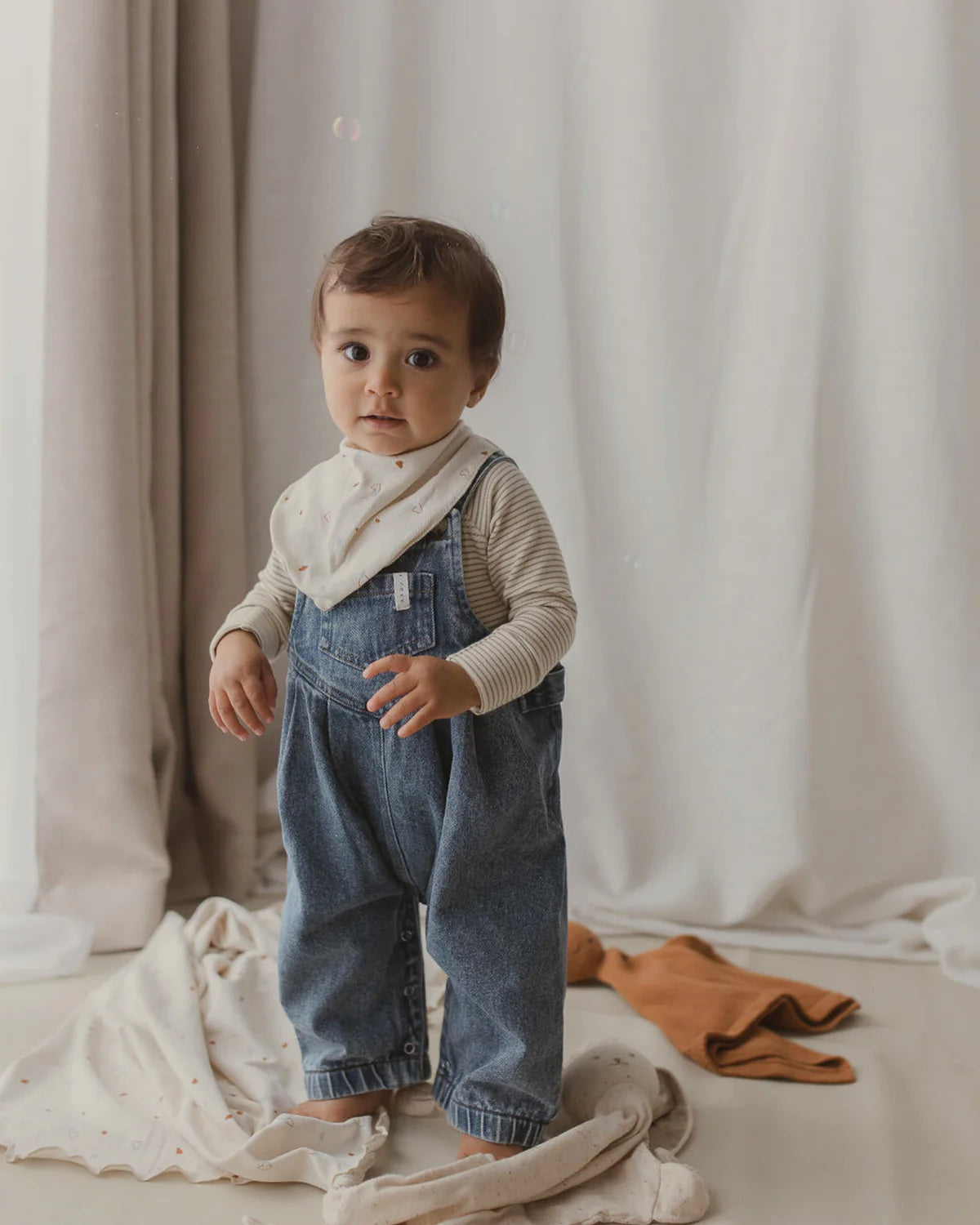 A toddler with short brown hair is standing indoors on a white blanket. The child is wearing SUSUKOSHI Denim Overalls in Sky Blue with adjustable shoulder straps over a beige long-sleeve shirt and a matching beige bib, all made from organic cotton. The background features a light gray curtain and some fabric casually placed on the floor.