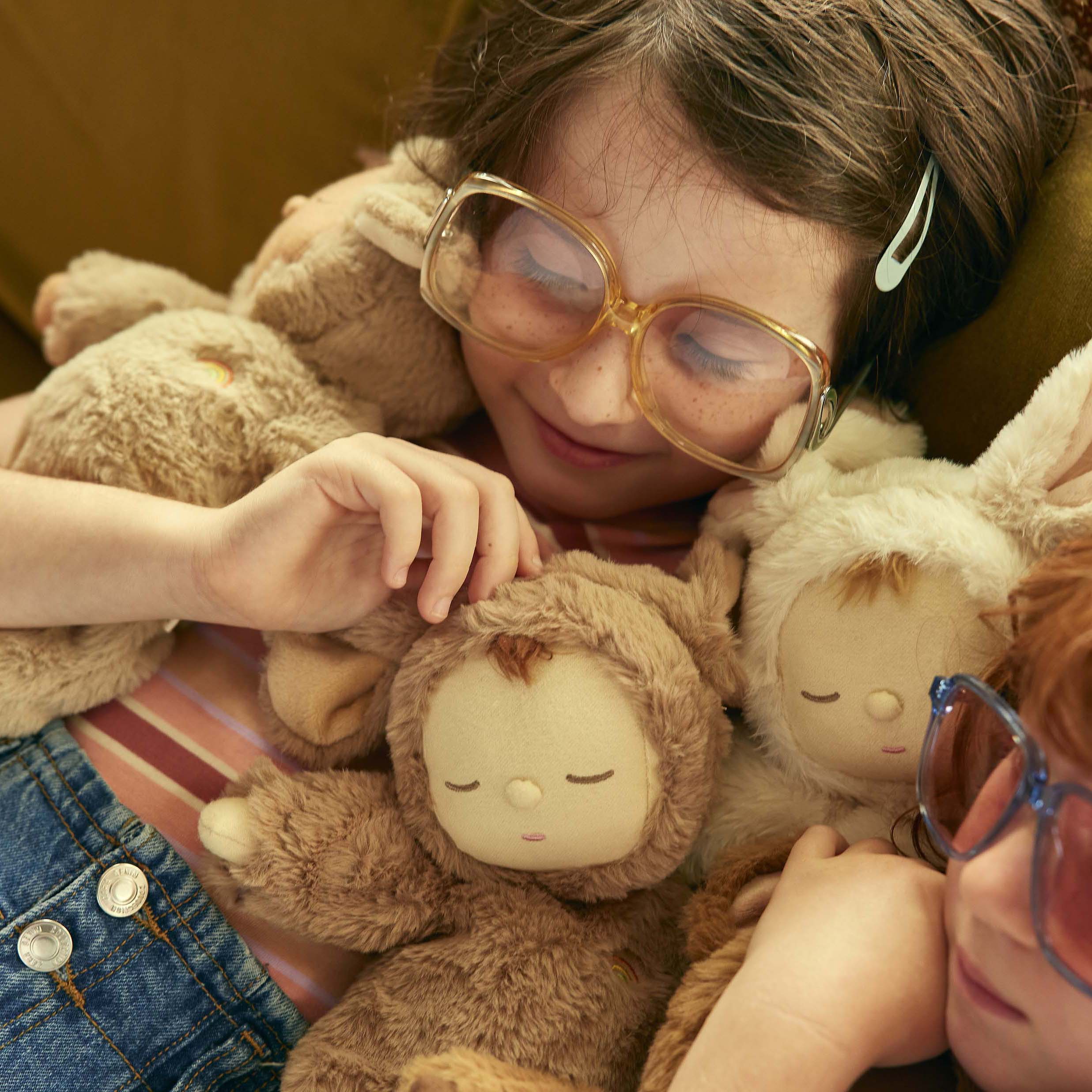 A child with glasses cuddles on a couch with multiple plush toys, including the Cozy Dinkum Lamby Pip by OLLI ELLA, smiling softly. Another child with glasses is partially visible, also surrounded by snuggly collectibles. Both children appear relaxed and content.