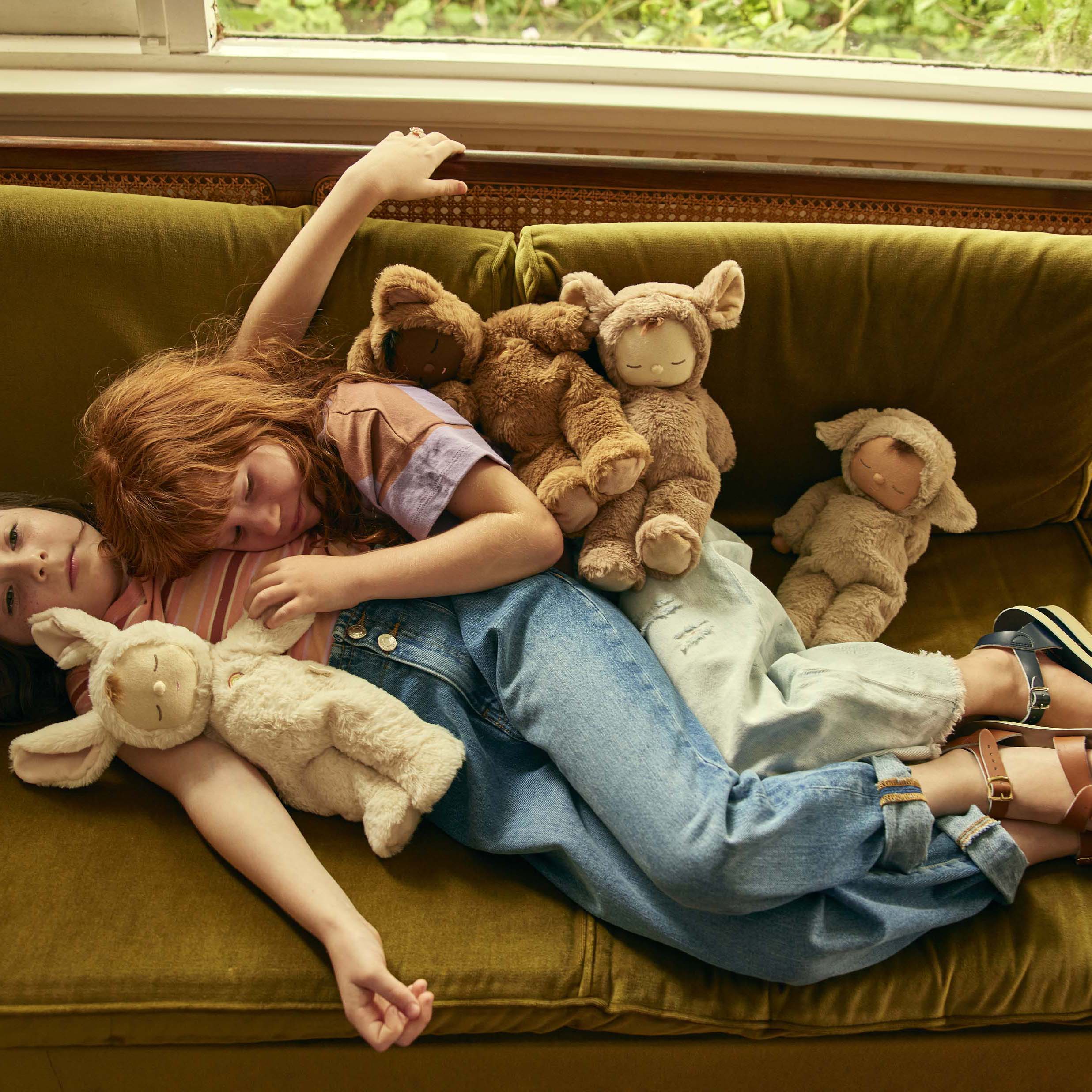 Two children are lying on a green couch, surrounded by several Cozy Dinkum Lamby Pip stuffed animals from OLLI ELLA. One child with red hair is resting on the other, who has dark hair. Both appear relaxed, enjoying a cozy moment together. There is a window with greenery in the background.