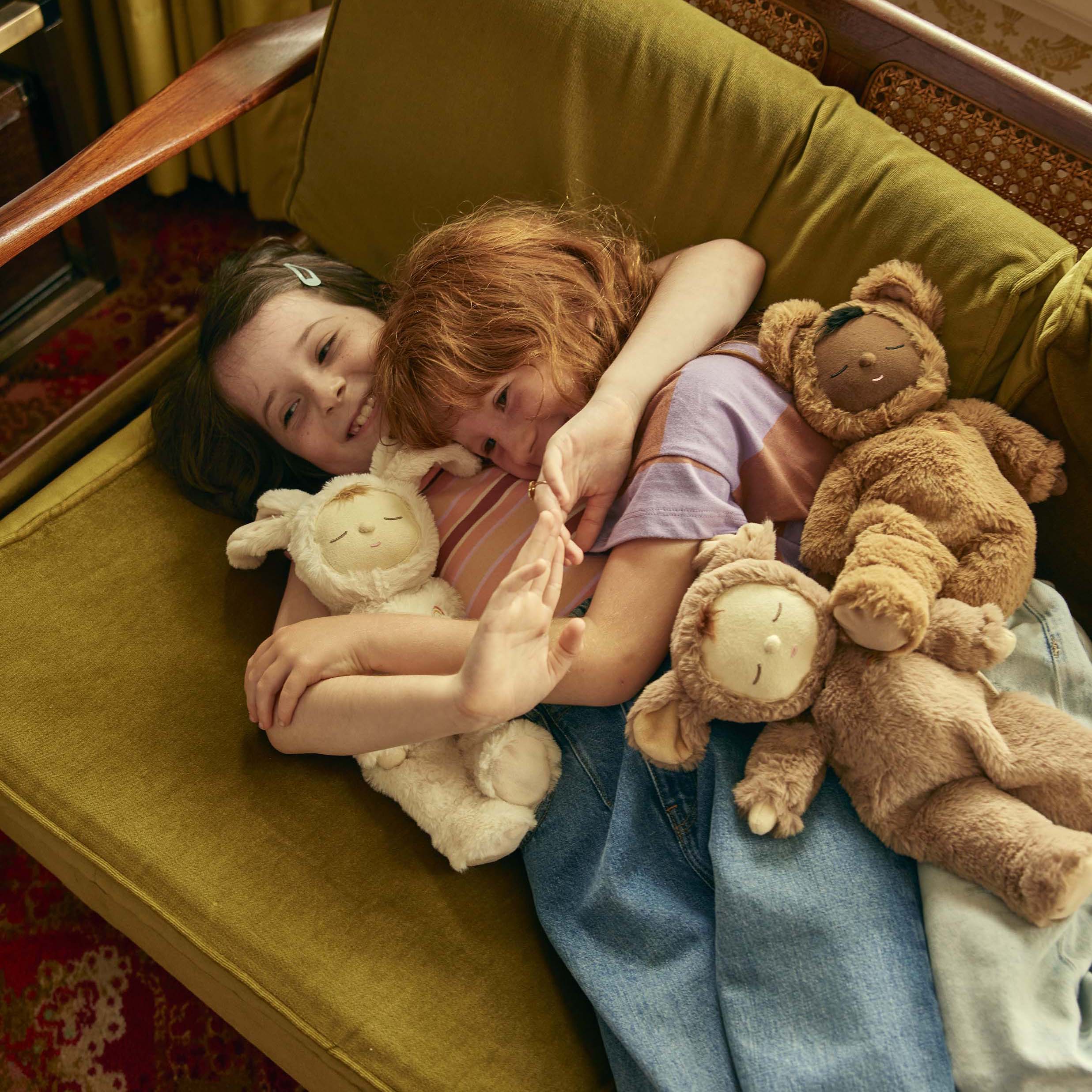 Two children lie on a green sofa, smiling and hugging each other, surrounded by plush teddy bears and a Cozy Dinkum Lamby Pip from OLLI ELLA. One child wears a pink striped shirt and has red hair, while the other wears a light-colored hair clip and a white shirt. The scene is cozy and cheerful.