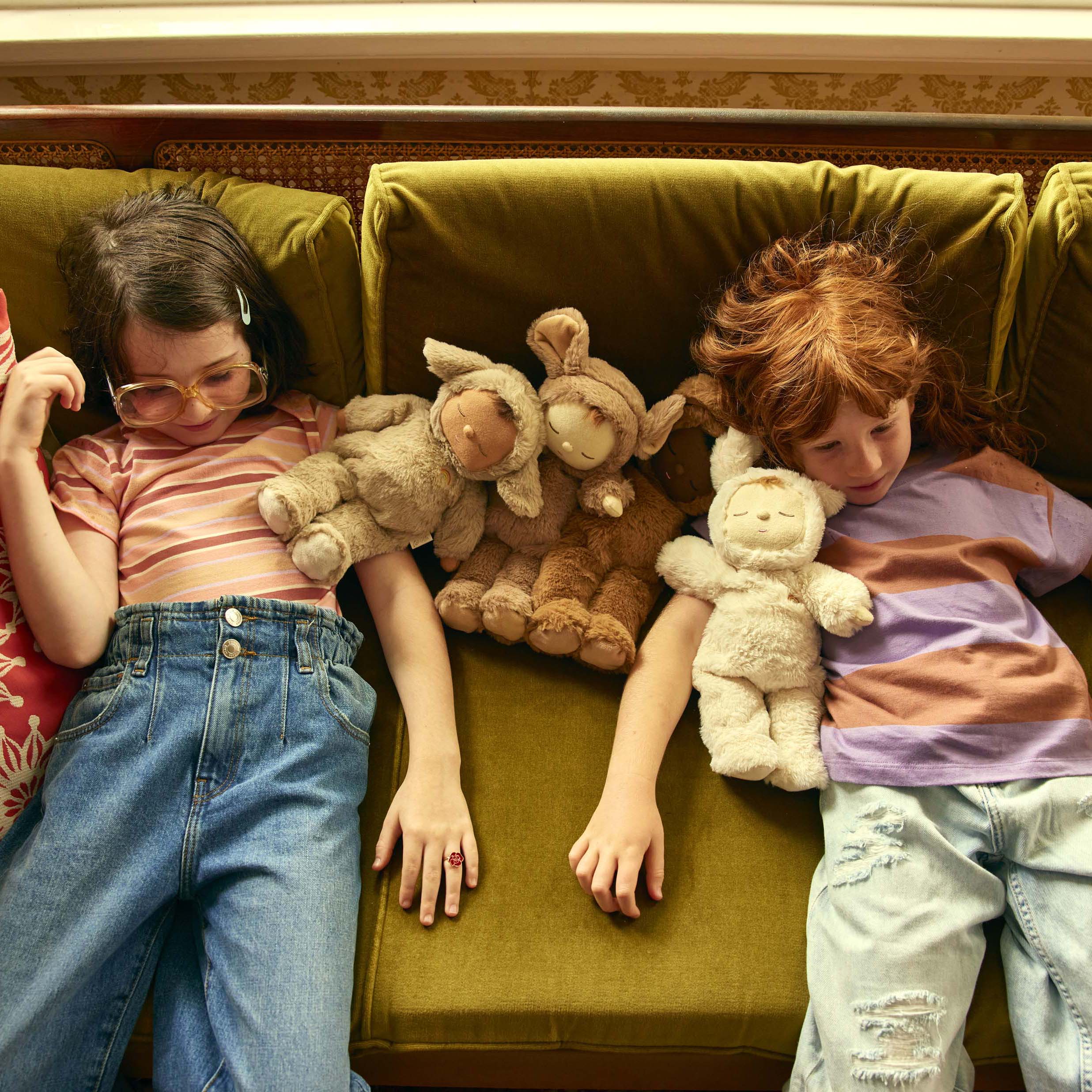 Two children lie on a green sofa, each holding a Cozy Dinkum Lamby Pip collectible by OLLI ELLA. One child wears glasses, a striped shirt, and blue jeans; the other has red hair, a striped shirt, and ripped jeans. They appear relaxed, surrounded by soft plush toys like their Cozy Dinkum Lamby Pips.