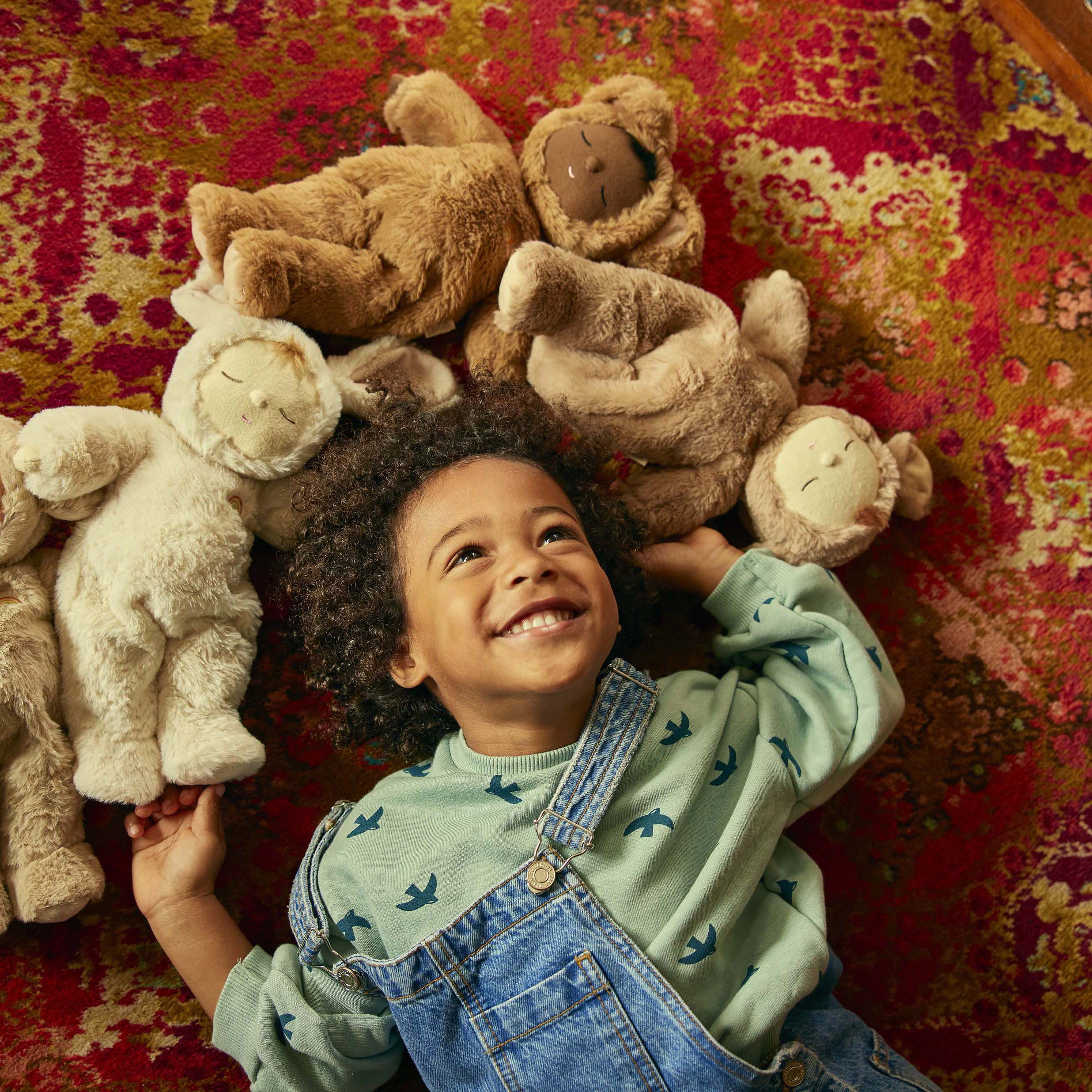 A young child with curly hair and wearing overalls lies on a colorful patterned rug, smiling up at the ceiling. They are surrounded by several plush toy animals, including bears, sheep, and a Cozy Dinkum Lamby Pip from OLLI ELLA, arranged in a circle around their head.