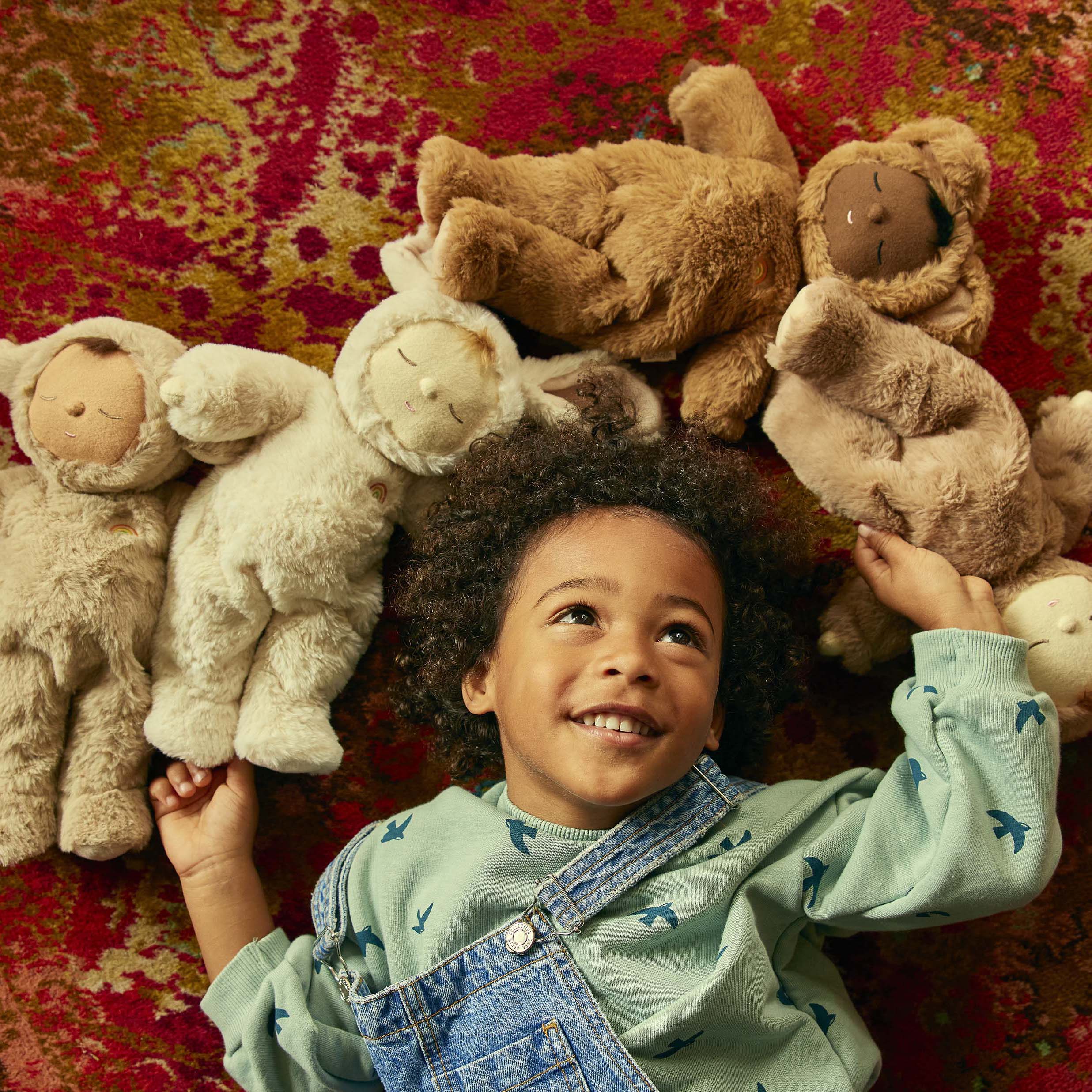 A child with curly hair lies on a red patterned rug, smiling and looking up. They are surrounded by six stuffed animals, including both teddy bears and the OLLI ELLA Cozy Dinkum Lamby Pip, positioned in a circle around their head. The child wears a light green shirt with bird patterns and denim overalls.