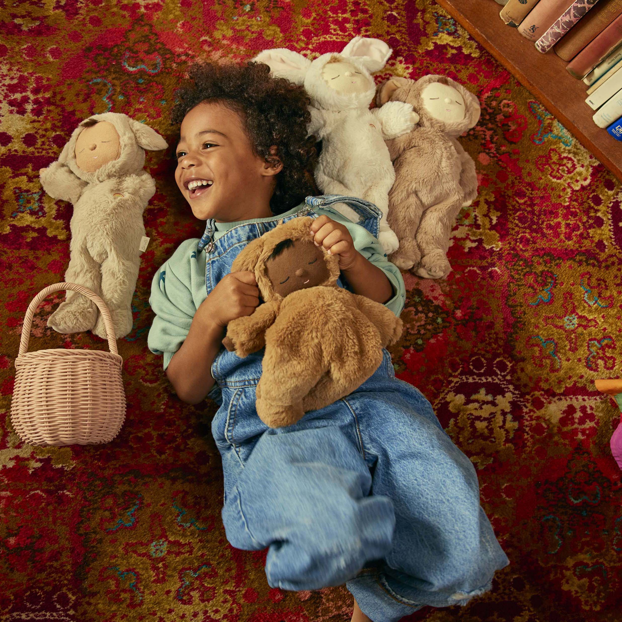 A child with curly hair and wearing overalls lies on a red patterned rug, surrounded by several plush toy animals, including the Cozy Dinkum Lamby Pip from OLLI ELLA. The child is smiling while holding one of the plush toys close. A small wicker basket is also nearby, and books are partially visible in the corner.