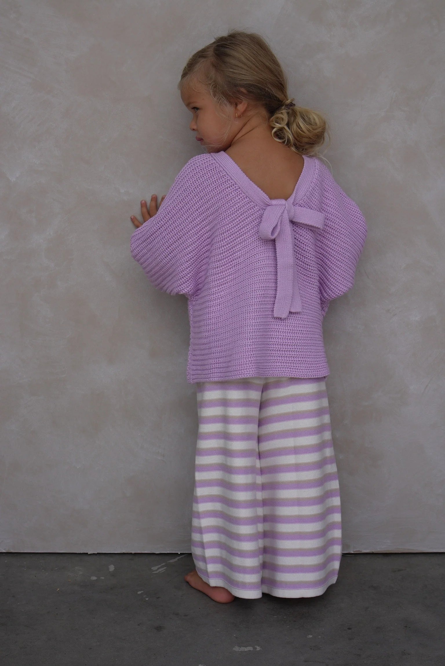 A young blonde child stands barefoot facing away, wearing a light purple knit sweater with a back bow and EN. THE LABEL's "Stripe Knit Pants Lilac," featuring white and purple stretchy fabric. The background is a plain wall.