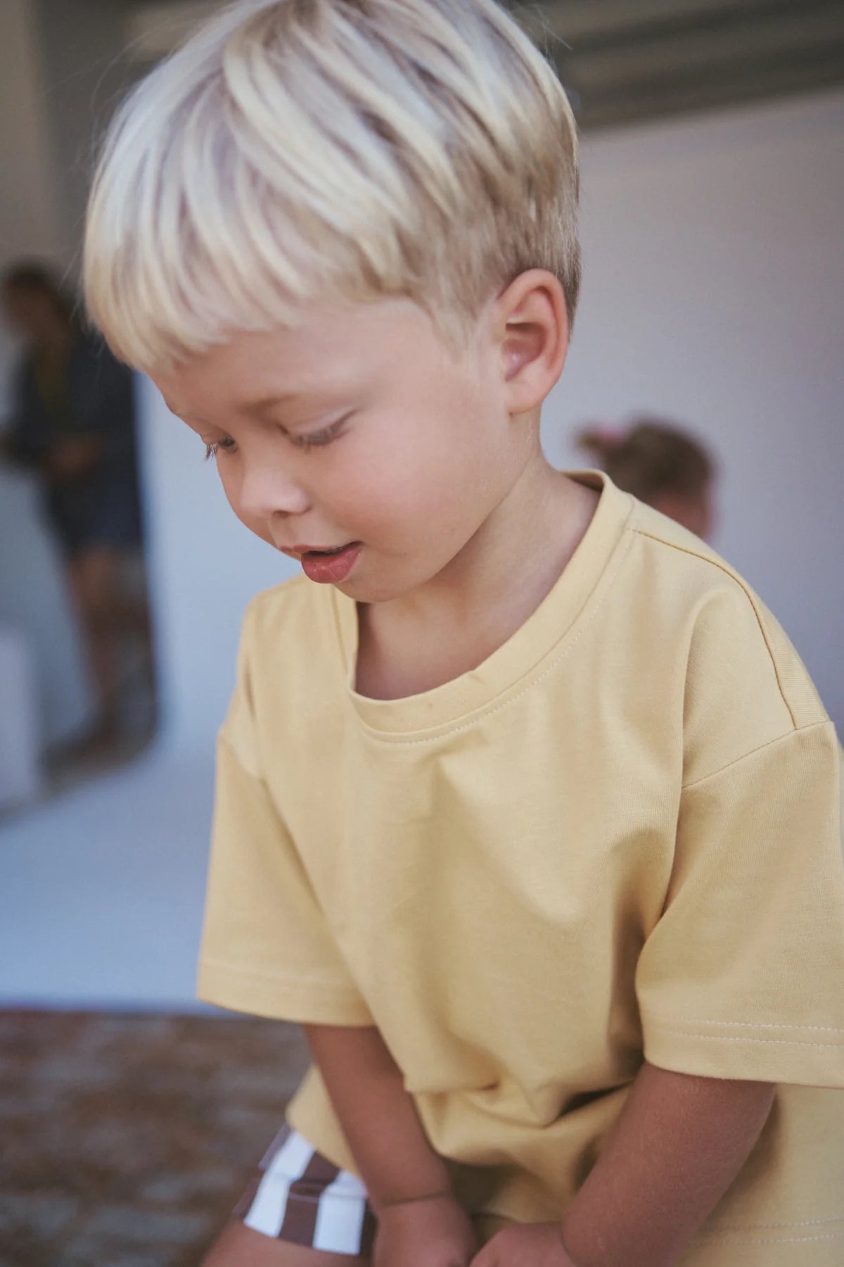 A young blonde child, wearing a Little Lou Tee Pomello from LITTLE THE LABEL, smiles slightly while looking down. The oversized tee is made from 100% cotton, and a softly blurred background shows another person in the distance.