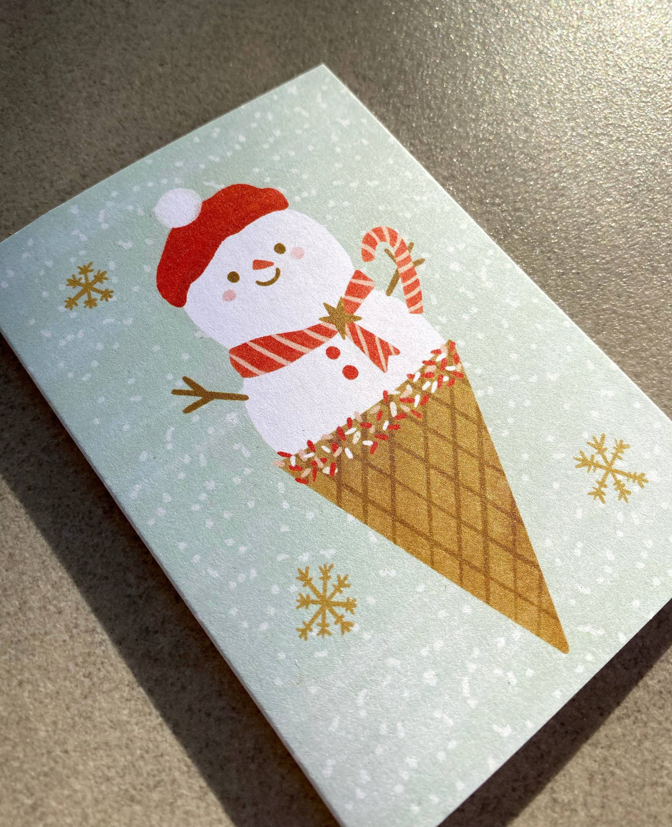 The Ms Snow Cone Mini Card by LAUREN SISSONS STUDIO is a delightful Christmas card made from recycled paper. It depicts an adorable snowman in a red hat emerging from an ice cream cone, holding a candy cane amid snowflakes on a light green background.