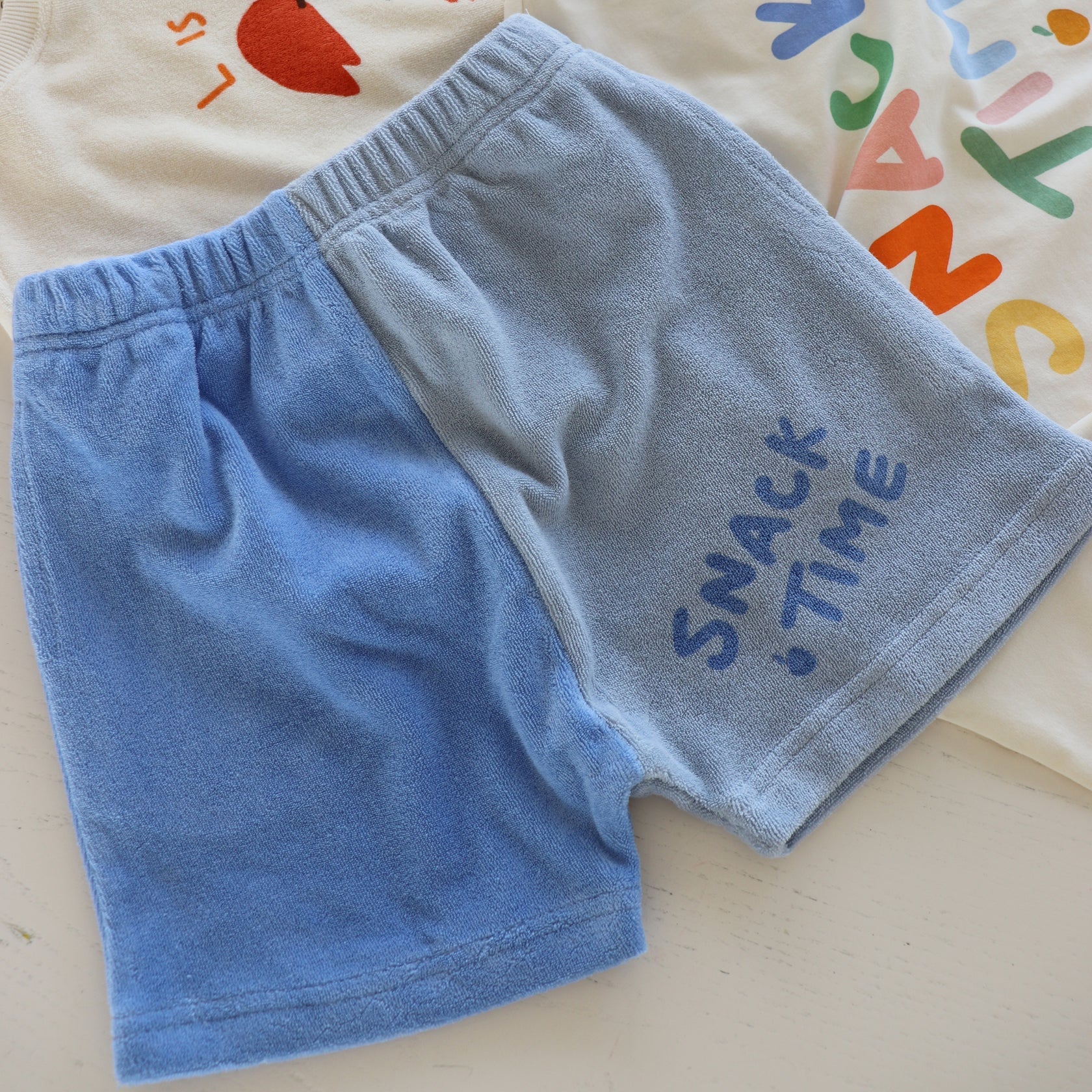 A pair of two-tone blue Play Lounge Shorts Pool from GOLDEN CHILDREN, with the words "SNACK TIME" printed on the right leg, lies on a white surface. A corner of a white garment with colorful lettering is partially visible in the background.