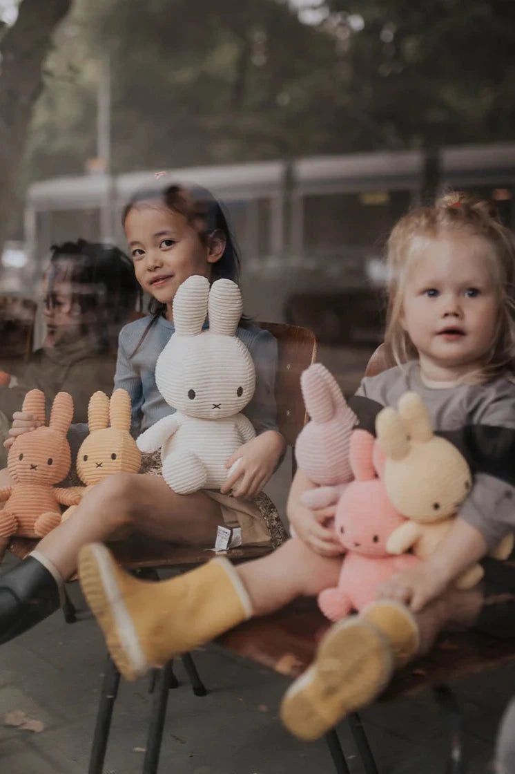 Two children sit behind a glass window, each holding plush bunnies in pastel colors. One child has dark hair and the other light, both cherishing their Miffy ECO Corduroy Off White toys from BON TON TOYS X MIFFY, filled with recycled PET material as they enjoy a cozy moment together.