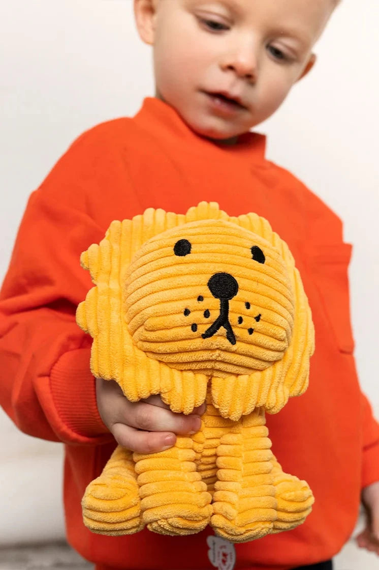 A child in an orange shirt holds a Lion Corduroy Yellow 24cm plush by BON TON TOYS X MIFFY. This soft, handcrafted toy is made from recycled polyester and has a simple, cute face against a light-colored background.