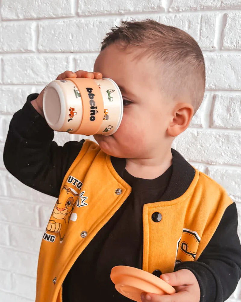 A young boy wearing a yellow and black jacket drinks from a small BBCINO Reusable BambinoCino Cup 240ml with cartoon truck designs, crafted from sustainably farmed bamboo. He's standing in front of a white brick wall.