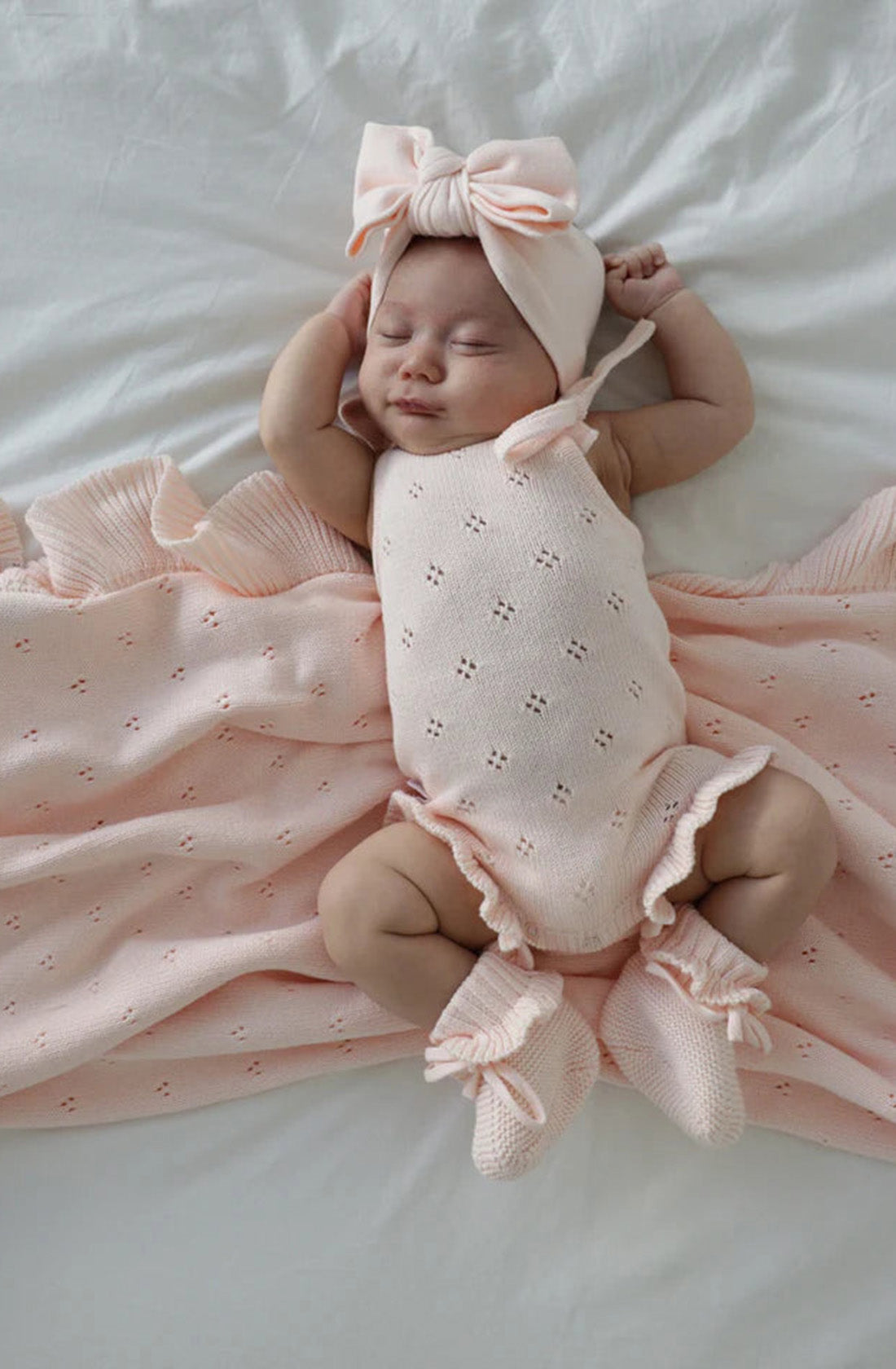 A sleeping baby wearing a light pink outfit from the Frill Bodysuit Primrose collection by ZIGGY LOU lies on a soft, white surface. The baby is dressed in a knitted frill bodysuit, matching booties, and a headband with a large bow. A light pink, ruffled blanket partially covers the baby. The baby has a peaceful expression.