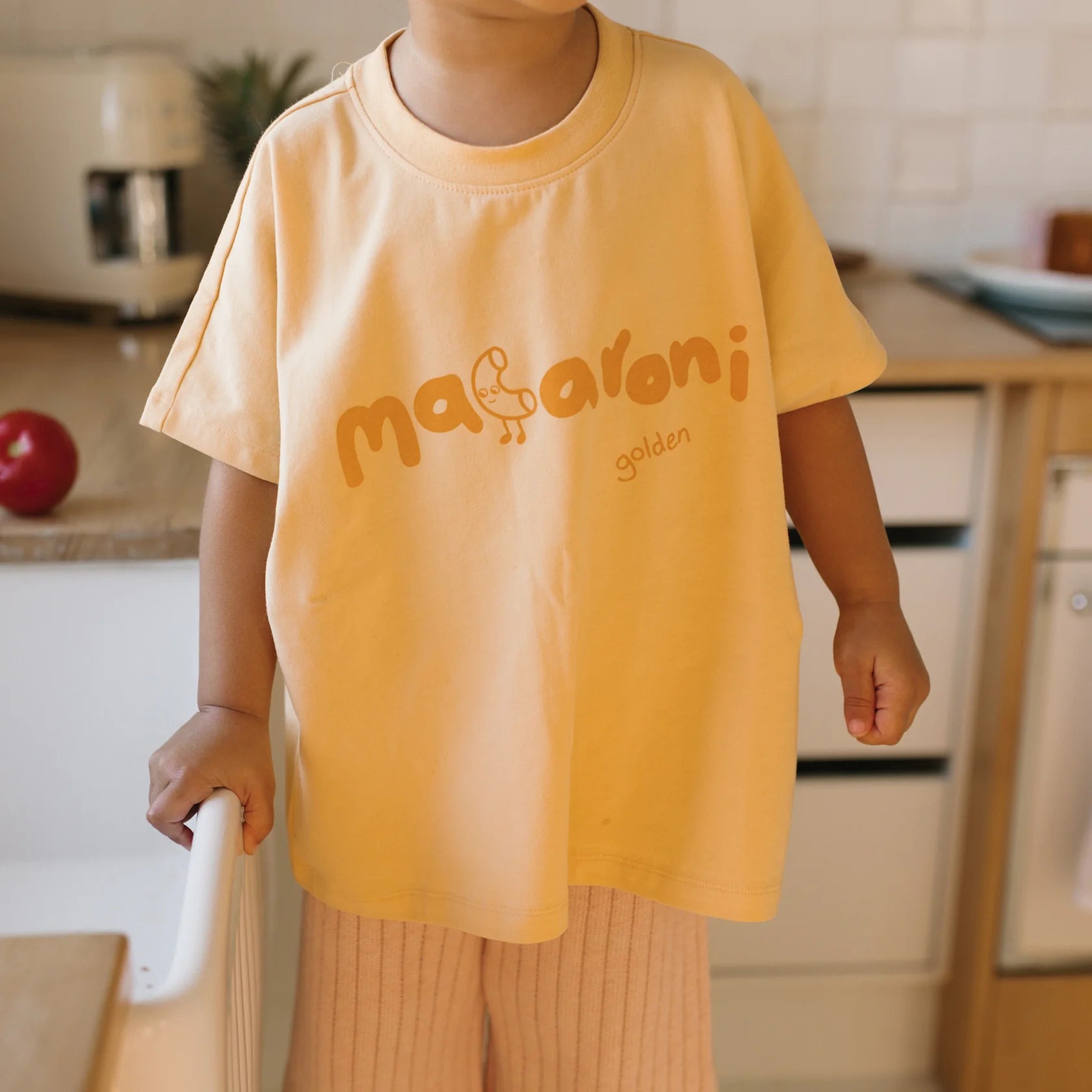 A child wearing an oversized Macaroni Tee Golden Butter by GOLDEN CHILDREN, made of soft stretch fabric with a small doodle of a duck printed on it, stands in a kitchen holding onto a white chair. A red apple and various kitchen items are visible in the background.