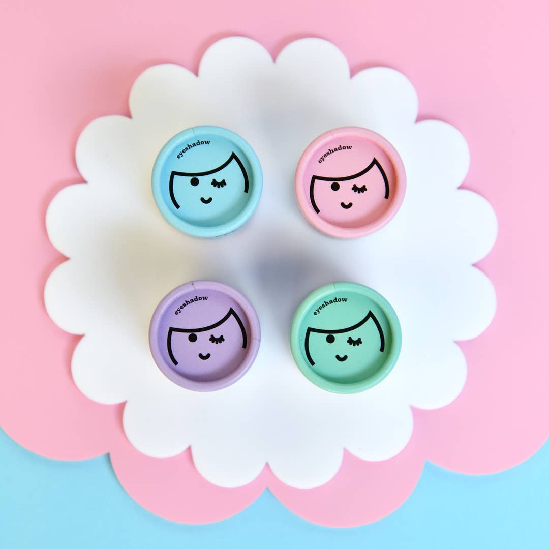 A flat lay photo displays four circular containers of no nasties kids Kids Eco Natural Pressed Eyeshadow & Blush, arranged on a scalloped white surface with a pink and blue background. Each container is adorned with a cute winking face design. The eyeshadow, created from natural ingredients, comes in purple, pink, blue, and green hues.