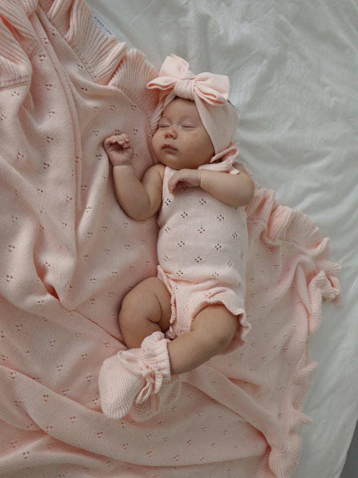 A baby peacefully sleeps on a light-colored blanket. The baby is dressed in a light pink Frill Bodysuit Primrose from ZIGGY LOU's Signature Collection, with a large matching bow on their head and cozy booties on their feet. The 100% cotton blanket has ruffled edges that complement the baby's attire.