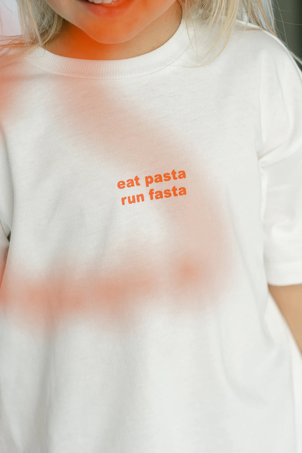 A close-up image of a person wearing the LENN LABEL Pasta Tee White made from soft cotton jersey. The kids tee features the words "eat pasta run fasta" printed in small orange letters on the front. The background is a blur of white and peach tones, with the person's face partially visible.