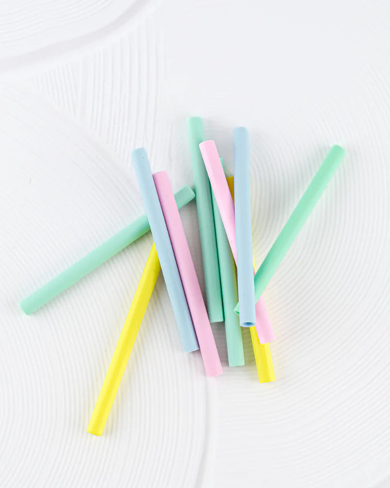 A collection of BPA-free, pastel-colored silicone straws from the BBCINO Replacement Silicone Straw Set Berry OR Dino—featuring shades of green, pink, blue, and yellow—are scattered randomly on a white textured surface. Eco-friendly and stylish, these straws add a touch of charm to any setting.