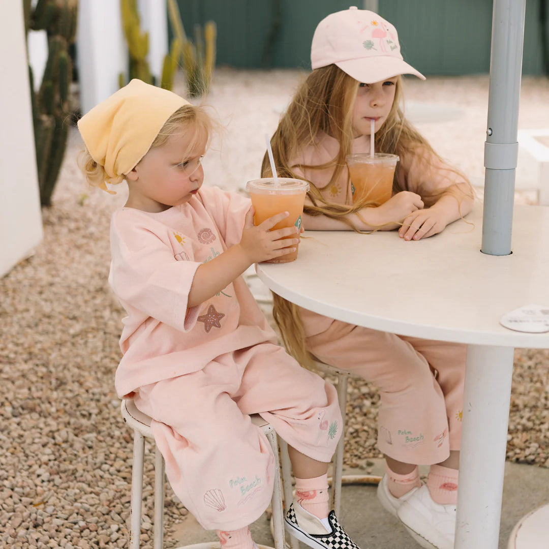 Two young children sit at a round outdoor table, both dressed in pastel pink outfits. One is wearing GOLDEN CHILDREN's Palm Beach Wide Leg Pants Terry Towel in Palm Flamingo Pink and sipping orange drinks from plastic cups with straws. One child has a hat on their head, while the other wears a yellow headband. The ground is covered with gravel.