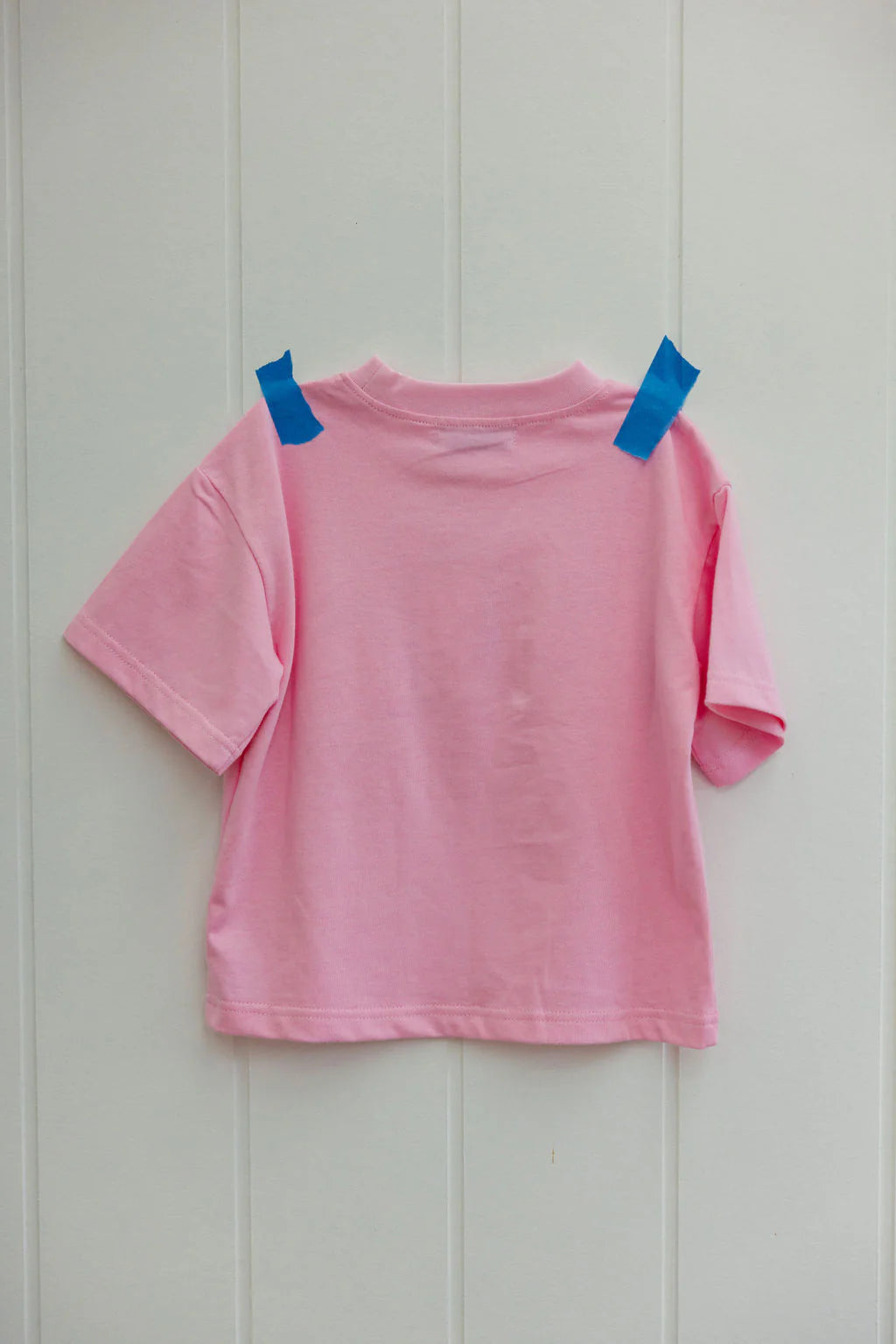 The Sunday Siblings ~ Il Sole Tee Pink/Red, an oversized piece by SUNDAY SIBLINGS, is affixed to a light gray wall with blue tape on its shoulders. Displayed backward, it highlights its 100% cotton material beautifully.