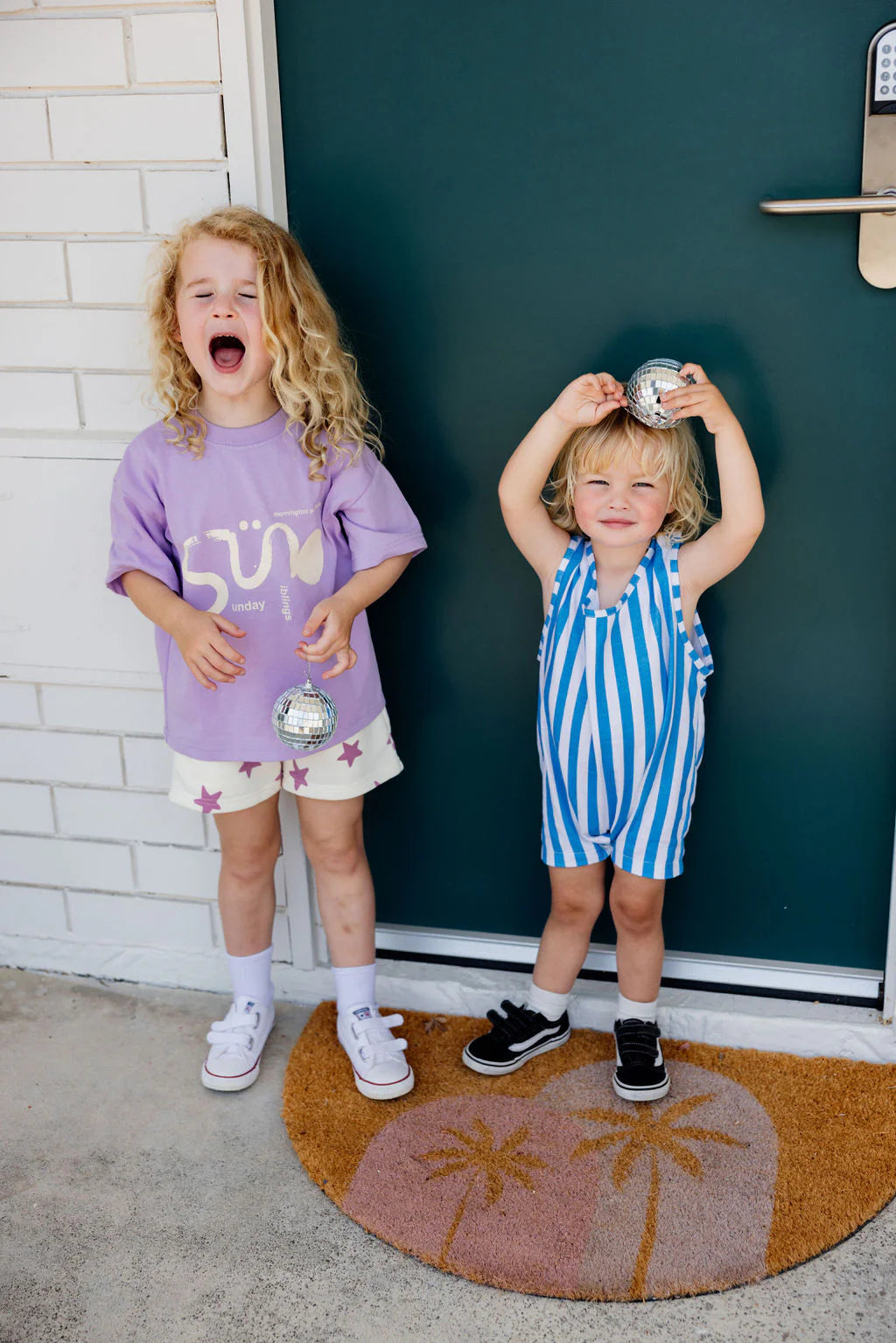 Two young kids stand on a doormat. One with curly hair in a purple Sunday Siblings Il Sole Tee looks excited, holding a small silver ball. The other, in an oversized fit blue and white cotton striped outfit, smiles holding a similar ball in front of a green door.