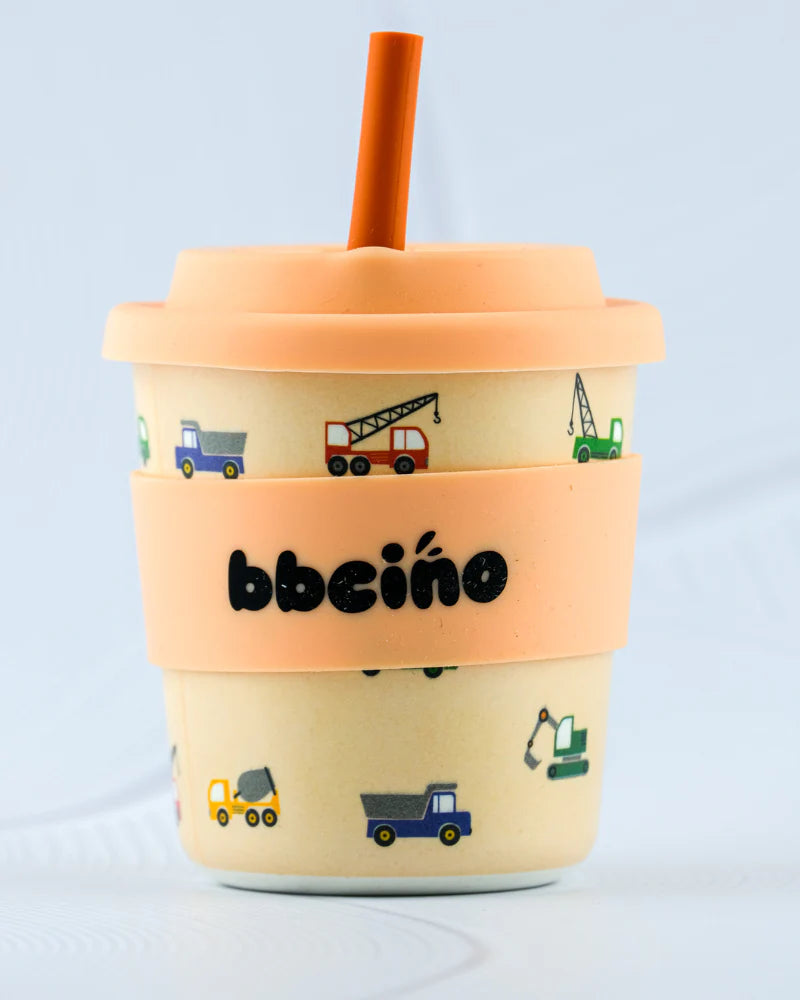 A sustainable 240ml sippy cup called the Reusable BambinoCino Cup by BBCINO features a cream exterior, peach lid, and orange straw. It is decorated with vibrant illustrations of construction vehicles such as trucks and cranes. The eco-friendly cup is accented with a peach band displaying the brand name "BBCINO" and highlights its production from sustainably farmed bamboo.