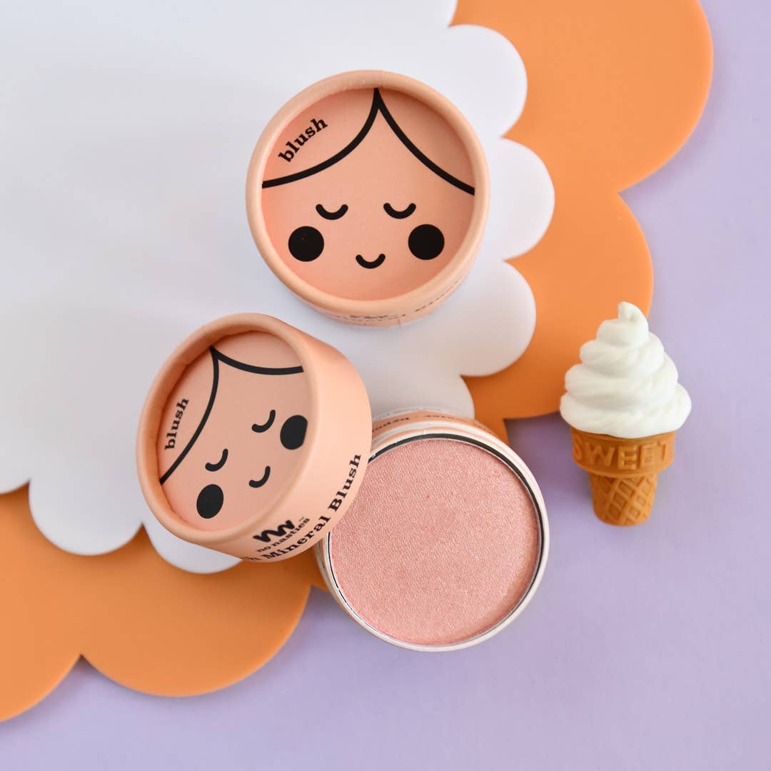 An open round compact of Kids Eco Natural Pressed Eyeshadow & Blush from no nasties kids reveals a shimmery pink powder made with natural ingredients. The lid, adorned with a cute face, rests beside it, displaying the word "blush" printed on the pink container. Nearby, an ice-cream-shaped decoration sits on a purple surface with an orange, scalloped-edge mat.