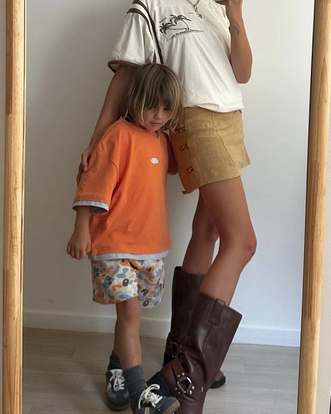 A woman and a child stand in front of a mirror. The woman rocks the trendy PRE-ORDER Double Layer Tee Paw Paw by FRANCO'S DAD, paired with tan shorts and knee-high brown boots. The child wears an oversized fit orange shirt and patterned shorts, both reflecting the playful style of pawpaw orange hues. They pose side by side indoors.