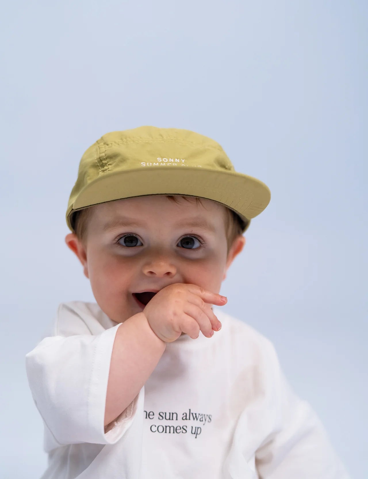 A baby wearing the Summer Club Cap in Sage by SONNY LABEL, which features an adjustable velcro strap, and a white shirt with text. The baby is touching their mouth and looking forward against a neutral background.